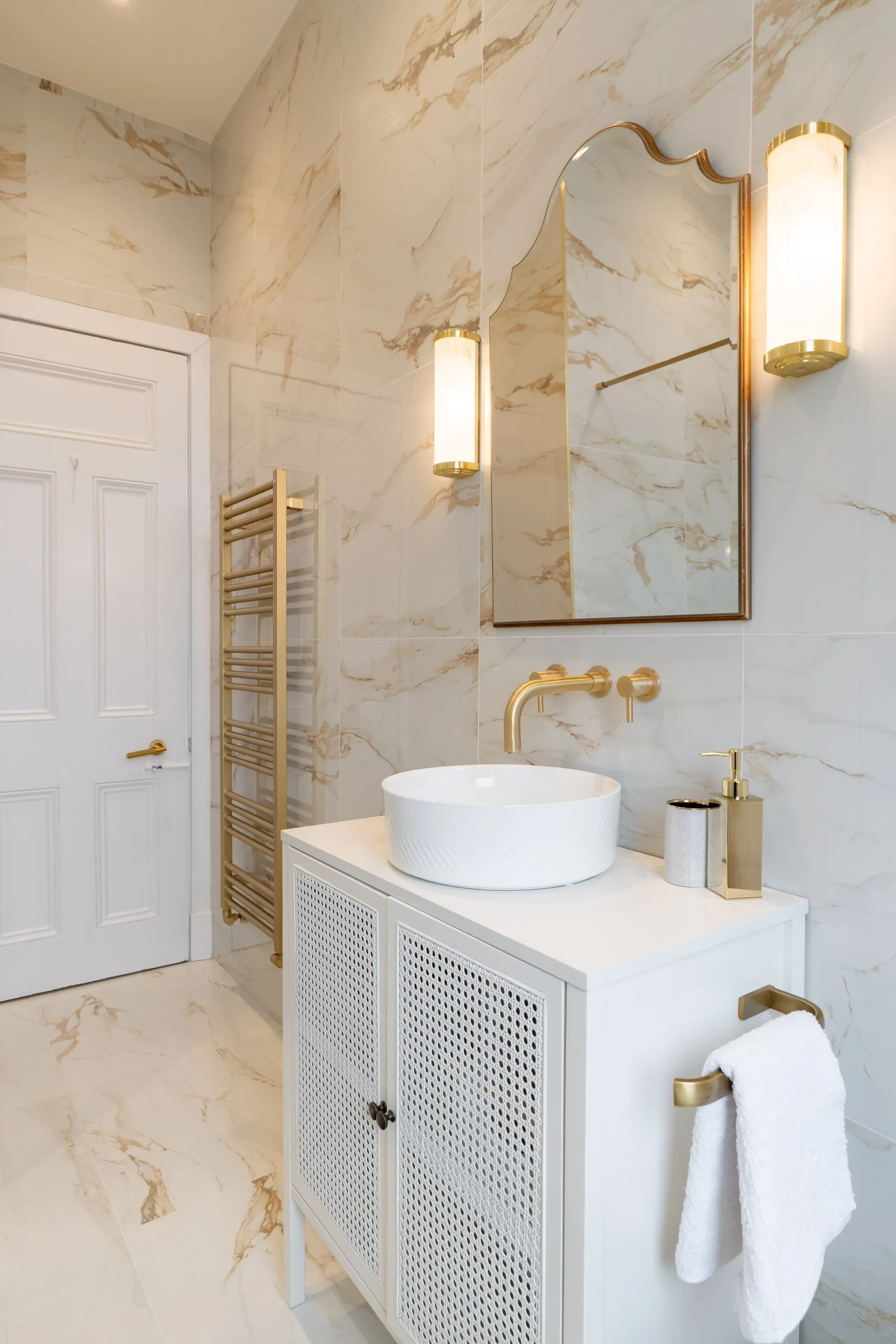 Modern bathroom with white marble walls and floor, gold accents, a white vanity with a vessel sink, a large mirror, wall-mounted lights, and a towel hanging on a gold towel rack.