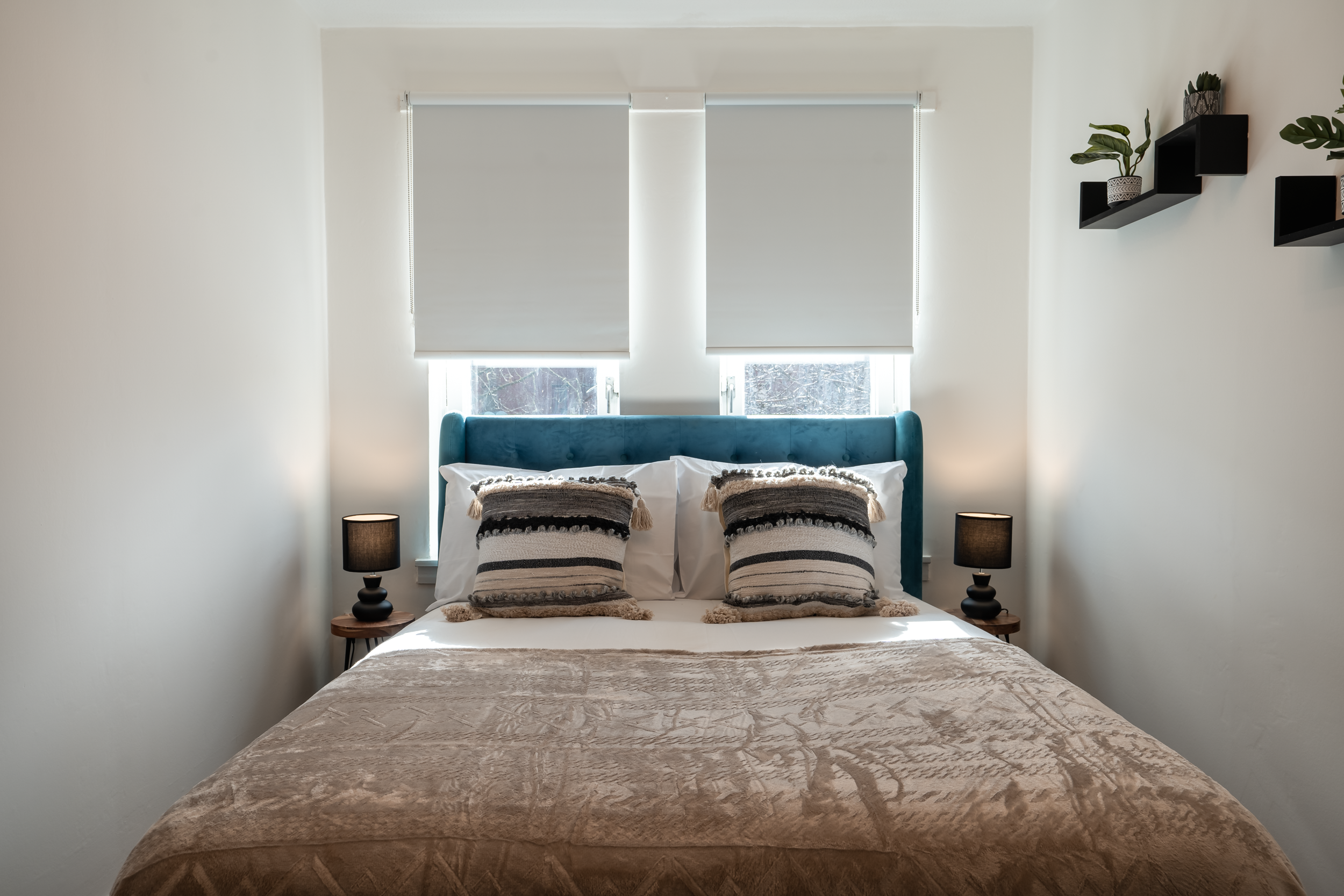 A well-made bed with a teal headboard, two striped throw pillows, and a beige blanket, flanked by two black table lamps in a bright bedroom with white walls and window blinds, and decorative black shelves with plants on the right wall.
