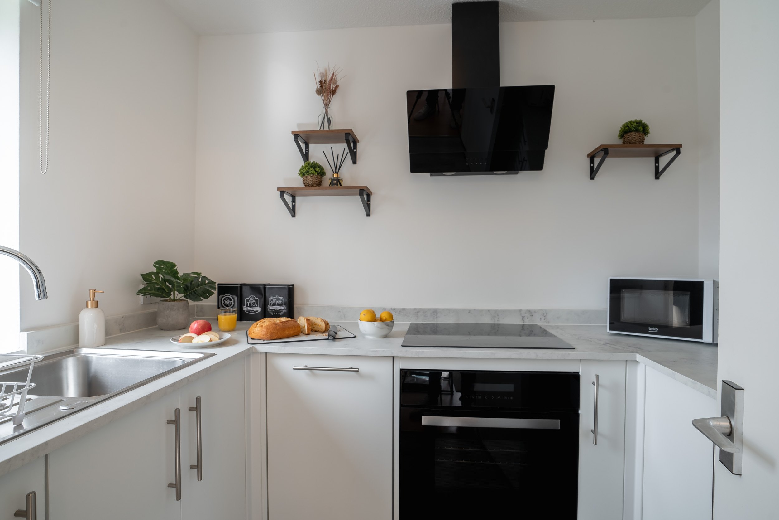 Modern kitchen with white cabinets, stainless steel sink, countertop with bread, fruit, and drinks, black stove, microwave, wall-mounted TV, and shelves with decorative plants and vases.