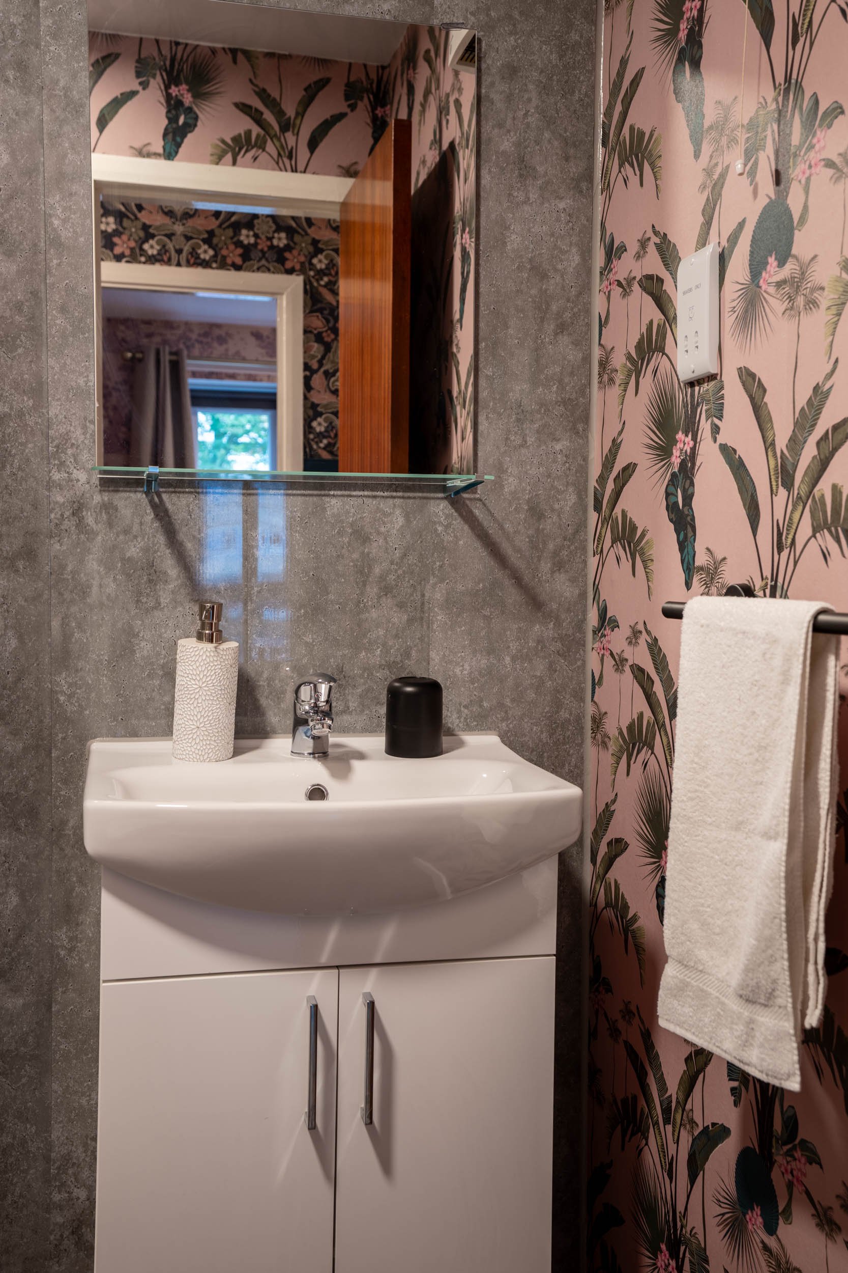 A small bathroom sink with a mirror, soap dispenser, black container, and a white cabinet below. There is a towel on a black towel bar and pink tropical wallpaper with green leaves and flowers on the walls.