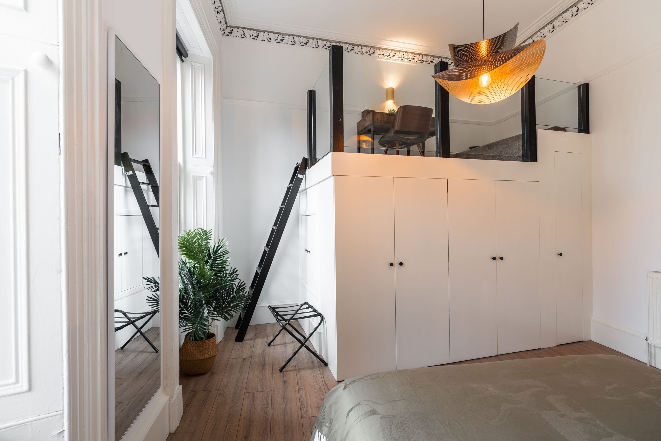 Interior of a bedroom with a white built-in wardrobe, a potted plant, a full-length mirror, and a black ladder. Upstairs area with a desk, chair, and modern pendant light.