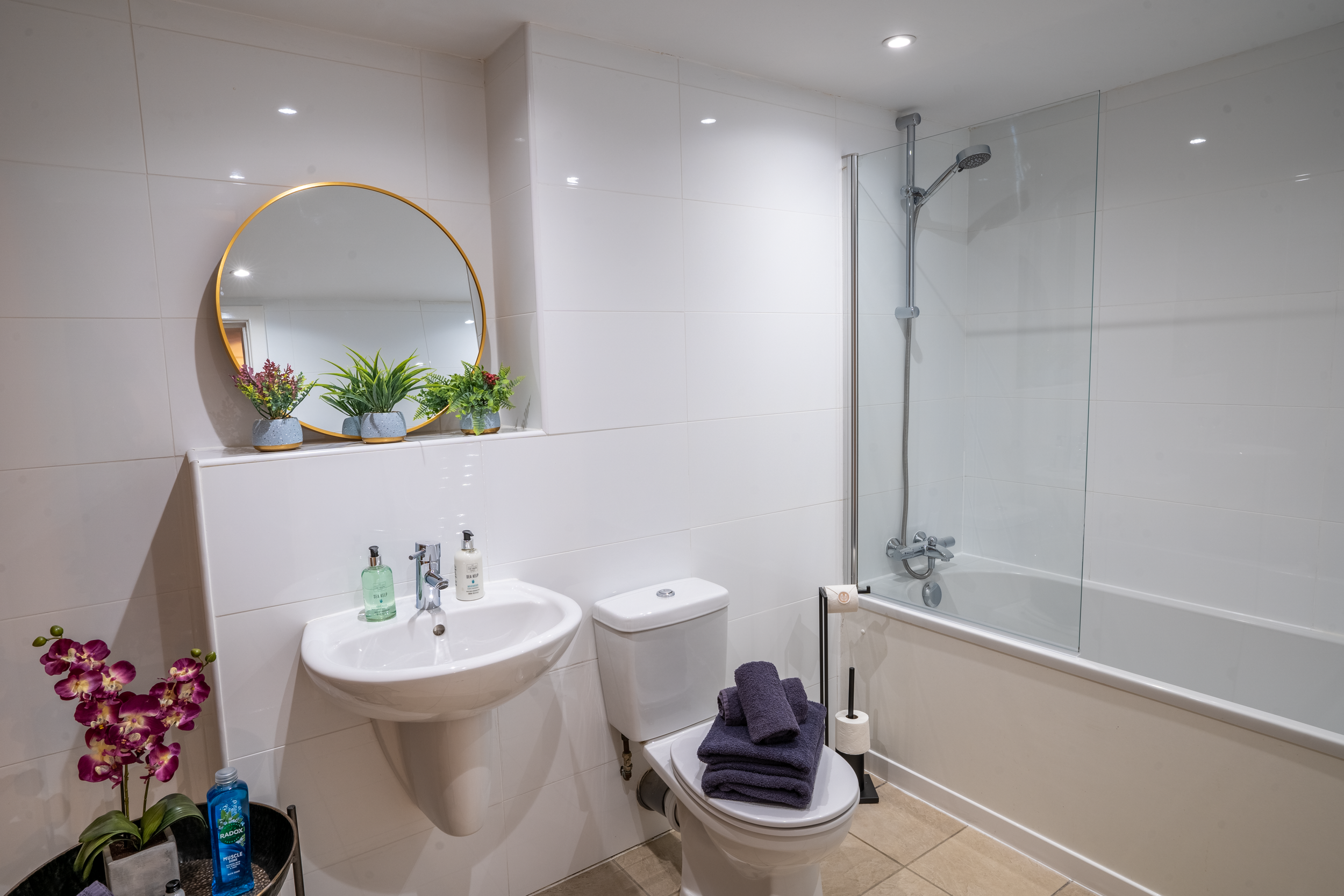Clean modern bathroom with white tiled walls, a sink with soap and sanitizer, a toilet with folded towels on the lid, a small black plant stand with pink orchids and personal care items, a wall mirror with three potted plants underneath, a bathtub wi