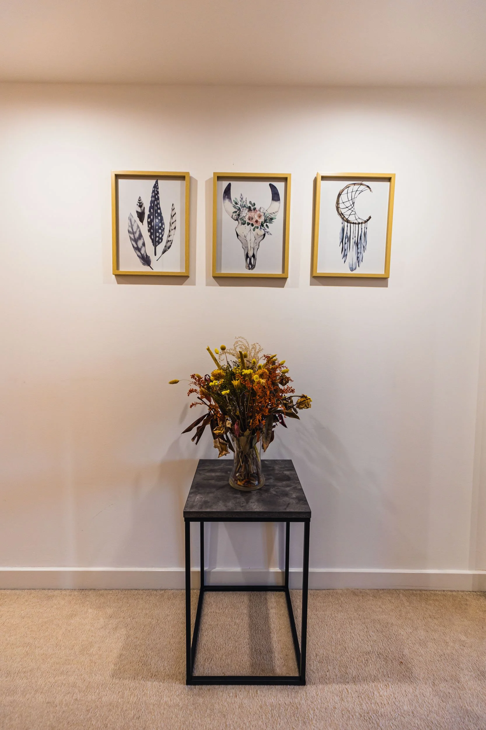 A wall with three framed art prints depicting feathers, a decorated animal skull, and a dreamcatcher. A small table with black metal legs holds a bouquet of dried flowers in a glass vase.