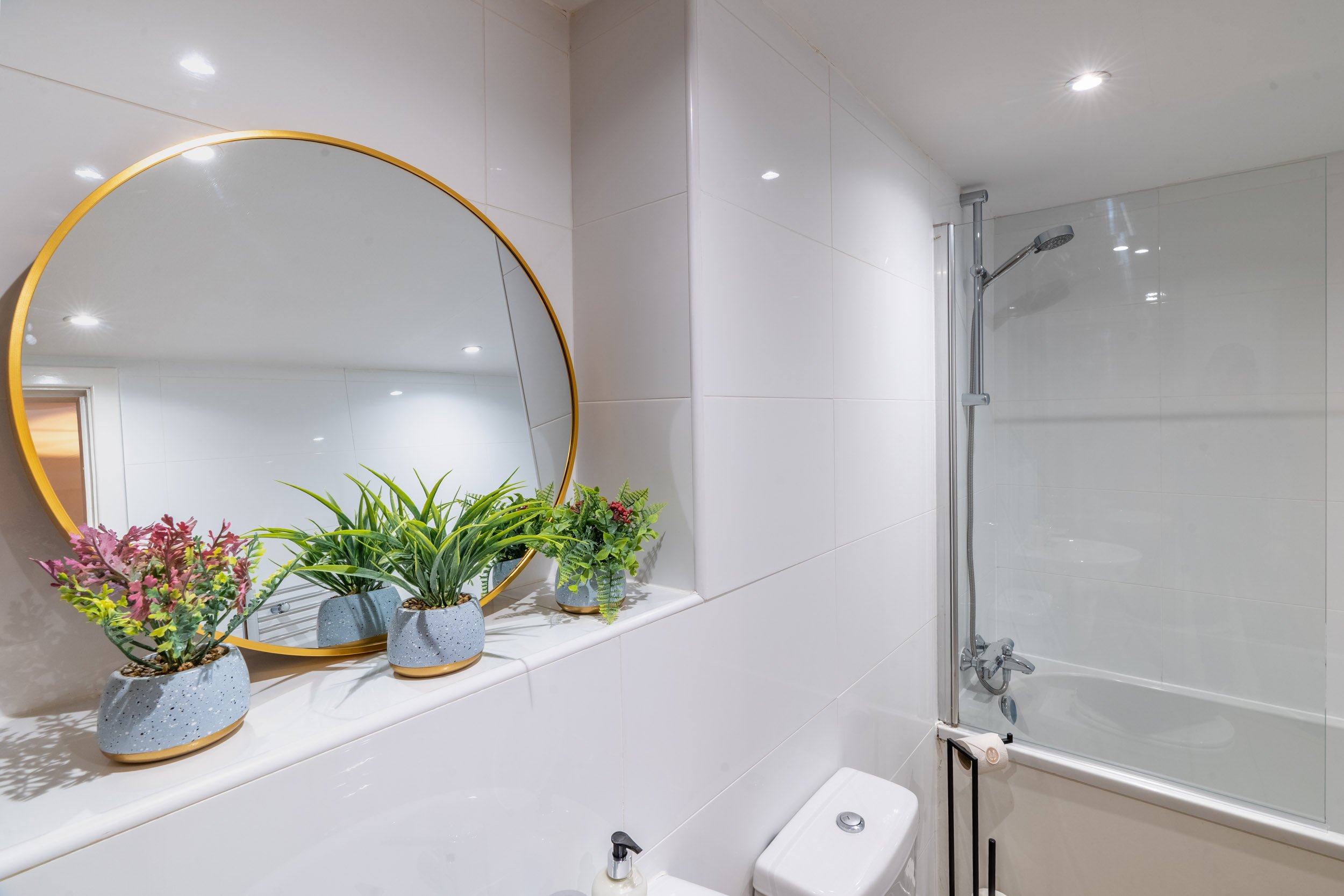 Bathroom with white tiled walls, a round mirror with a gold frame, potted plants on a shelf, a toilet, and a shower with a glass door and a handheld showerhead.