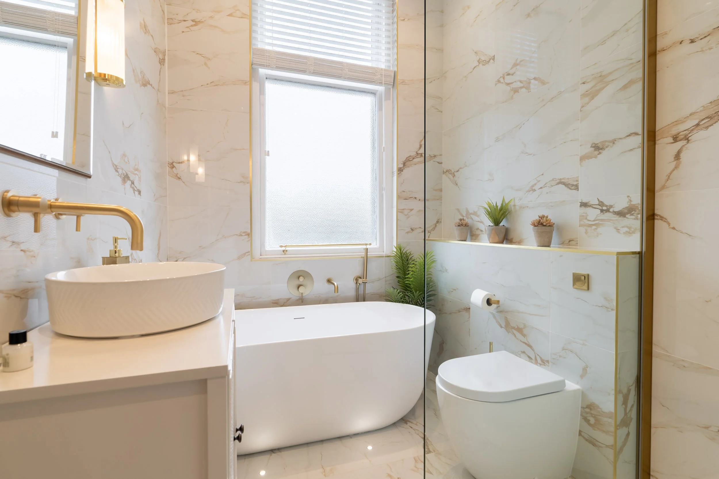 Modern bathroom with a white bathtub, a white vanity with a vessel sink, gold fixtures, a toilet, and decorative plants on a ledge. The walls and floor are covered in cream-colored marble tiles with gold veining. There is a frosted window with blinds