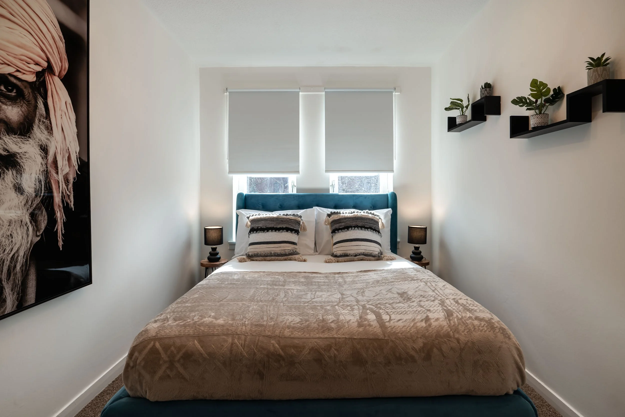 A neatly made bed with a teal upholstered headboard, two pillows with striped covers, and a beige blanket, flanked by two black nightstands with small black lamps, in front of a window with closed white blinds and a view of snow outside. There are bl
