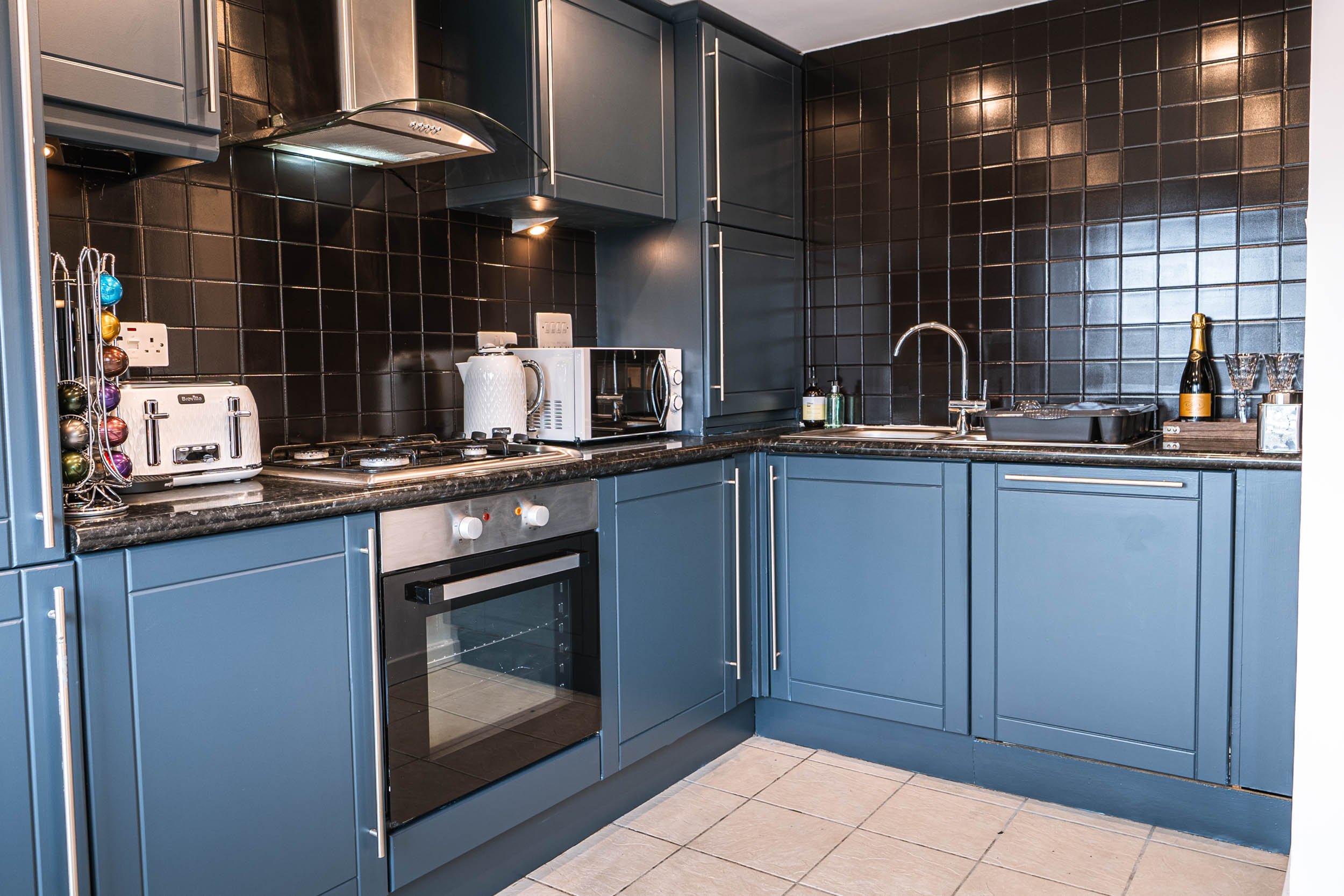 Kitchen with blue cabinets, black tiled backsplash, stainless steel appliances, and a beige tiled floor.