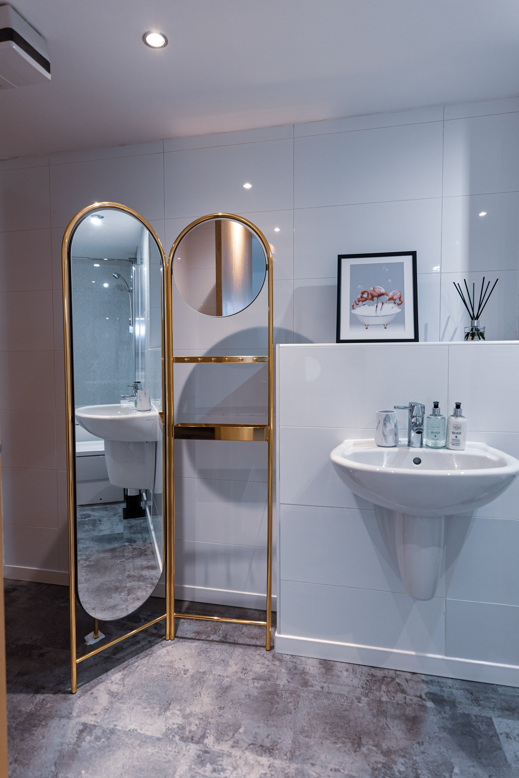 A modern bathroom with a gold-framed mirror and a small round mirror, a white wall-mounted sink with toiletries, a framed picture of flamingos, a reed diffuser, and a gold bookshelf divider