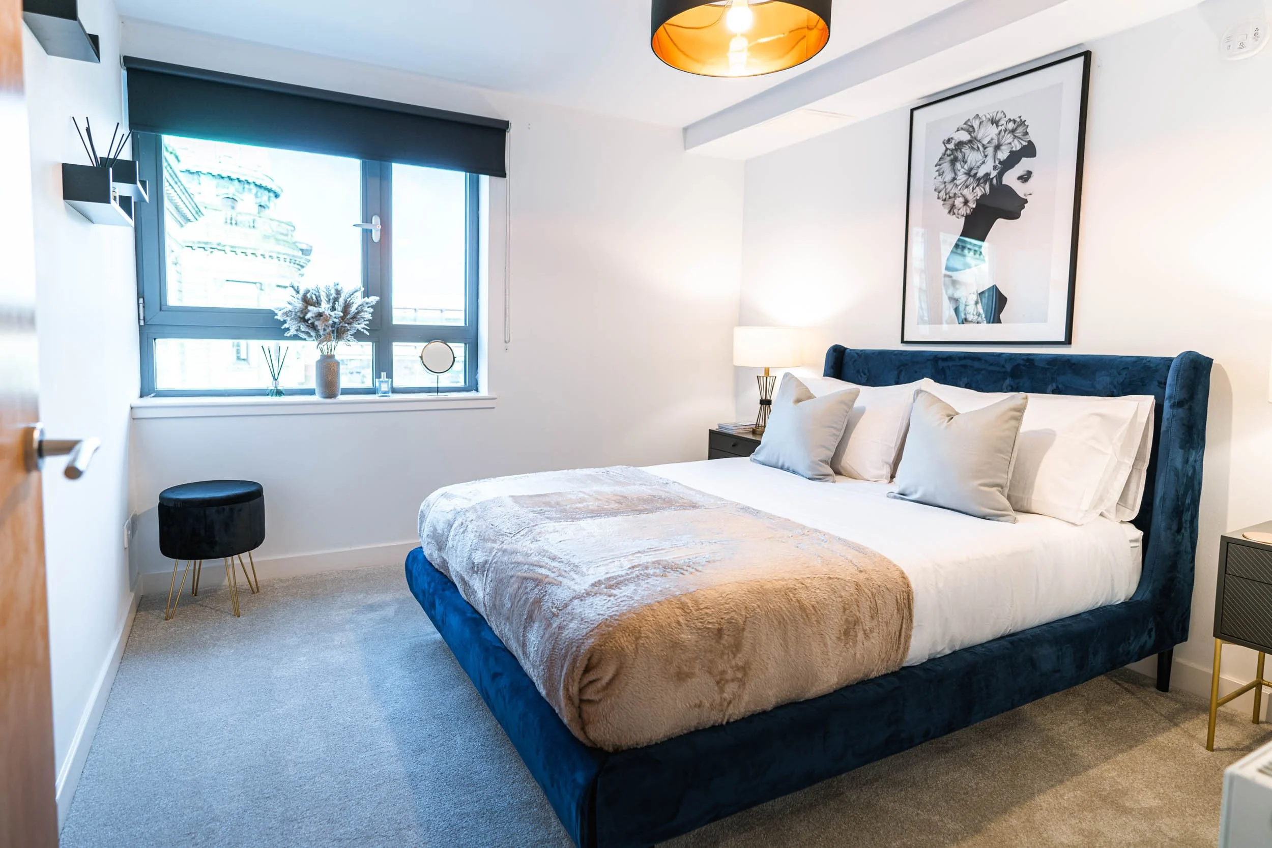 A modern bedroom with a large bed featuring a blue velvet headboard, white bedding, and beige and gray pillows. There's a framed black-and-white artwork on the wall above the bed. Nightstands with lamps are on each side, and a window with a dark blin