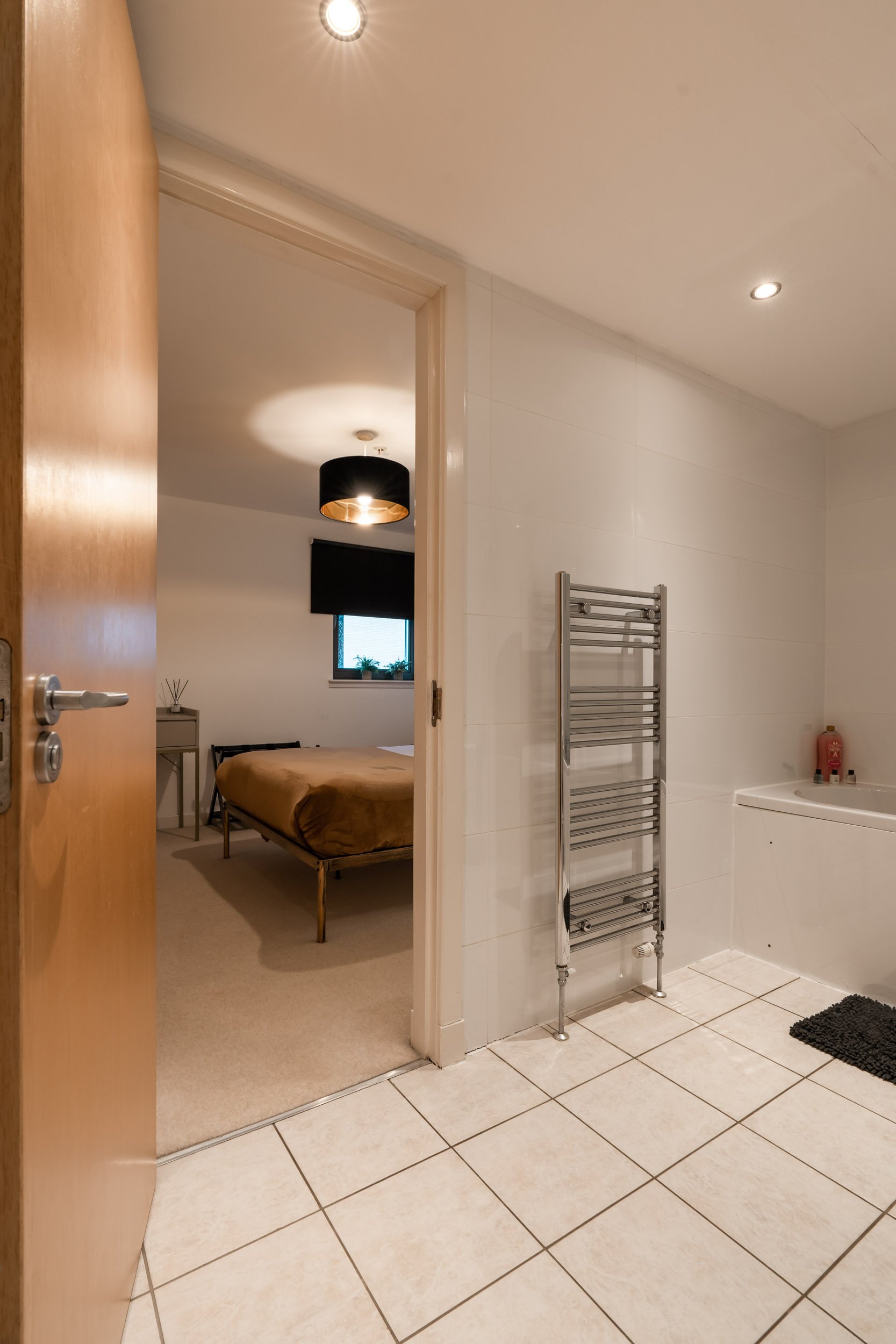 Bathroom with a heated towel rack, bathtub, black bathroom rug, and partially open door revealing a bedroom with a bed, window, black curtain, and ceiling light.