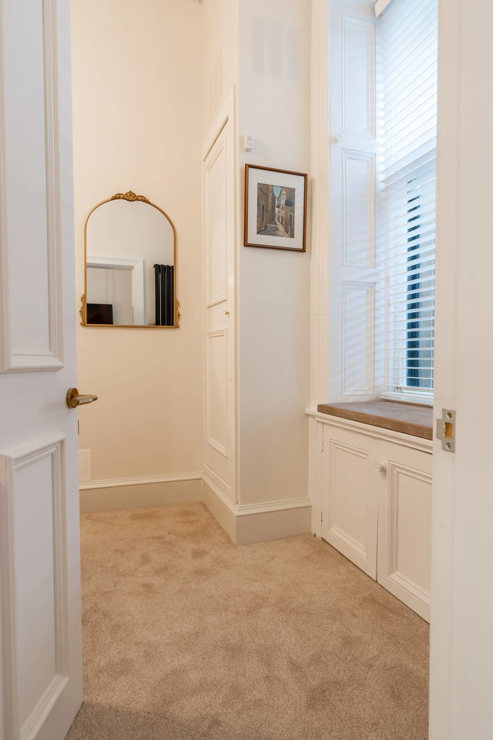 A small hall or entryway with a beige carpet, white walls, a window with white blinds, a framed picture, a decorative mirror, and built-in cabinets.