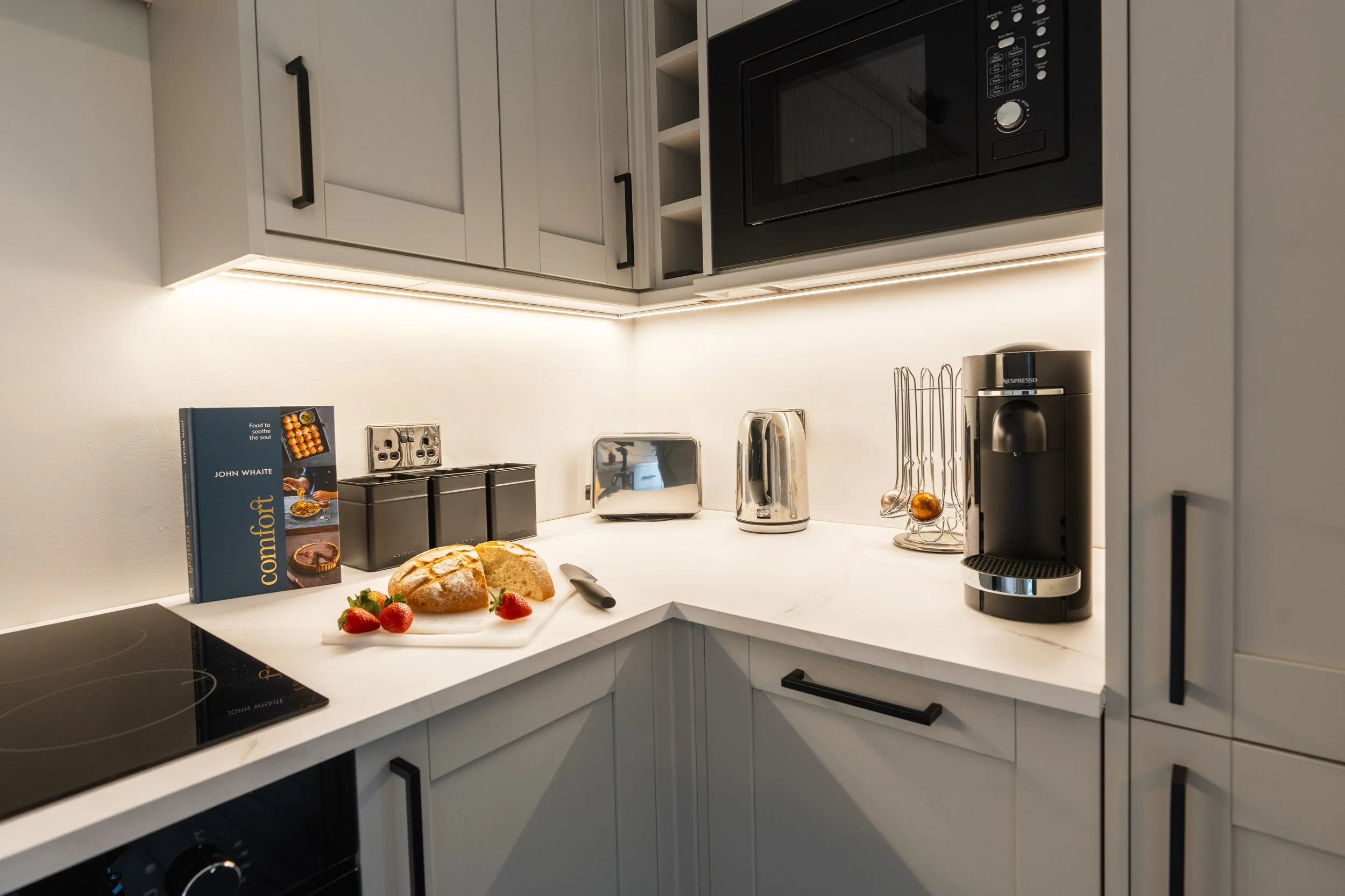Kitchen countertop with bread, strawberries, a knife, a cookbook, a toaster, a kettle, a Nespresso coffee machine, and utensils.