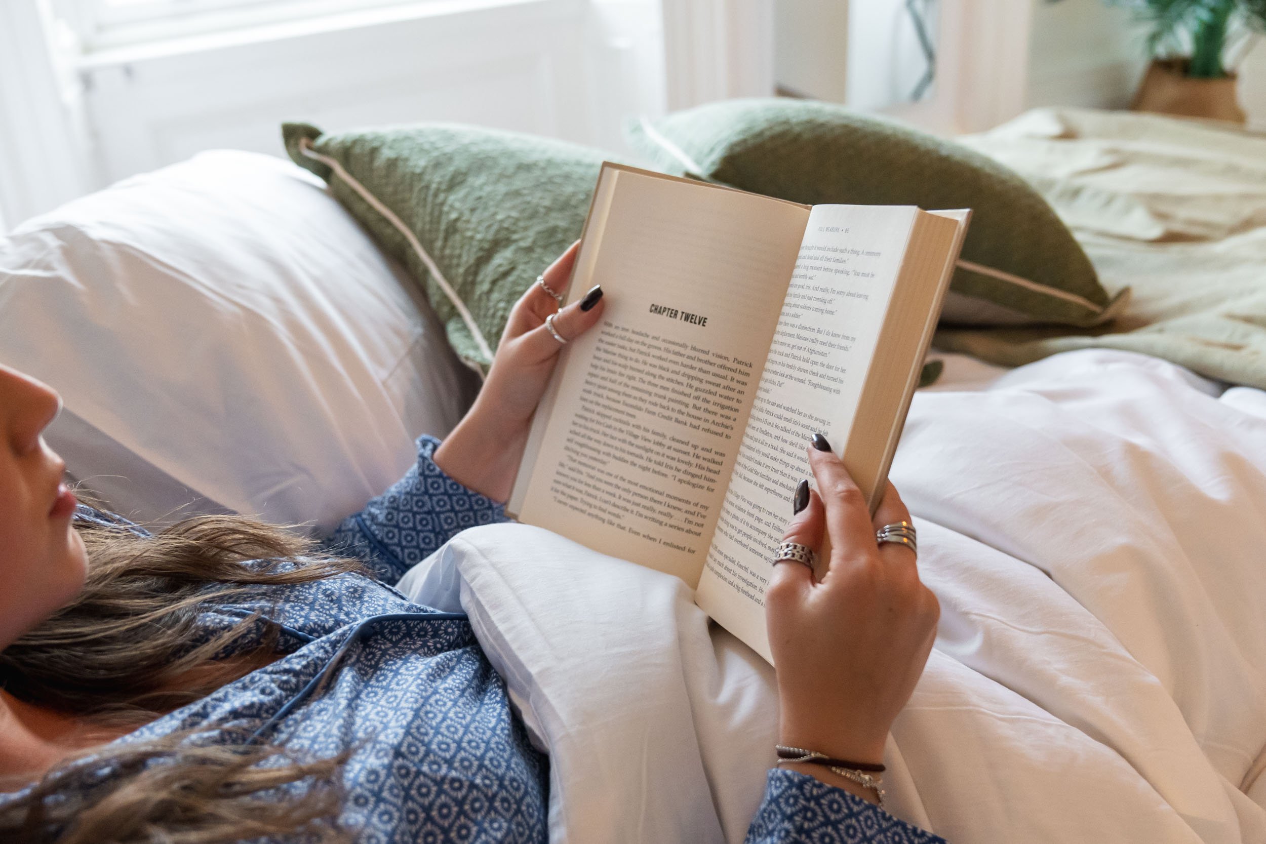 Person lying in bed reading a book, with pillows and blankets around them.