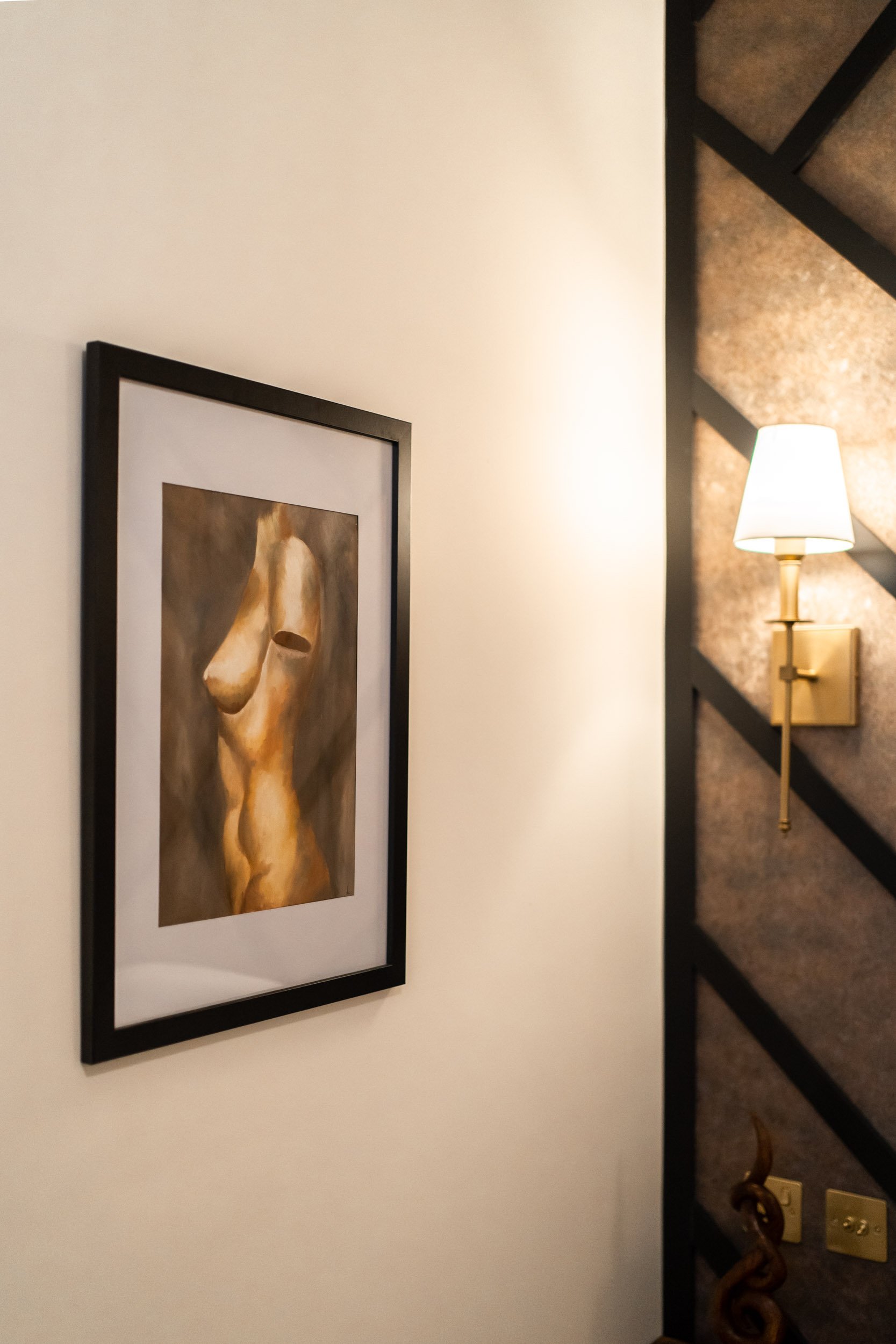 A framed painting of an abstract female torso on a white wall, with a wall sconce light nearby.