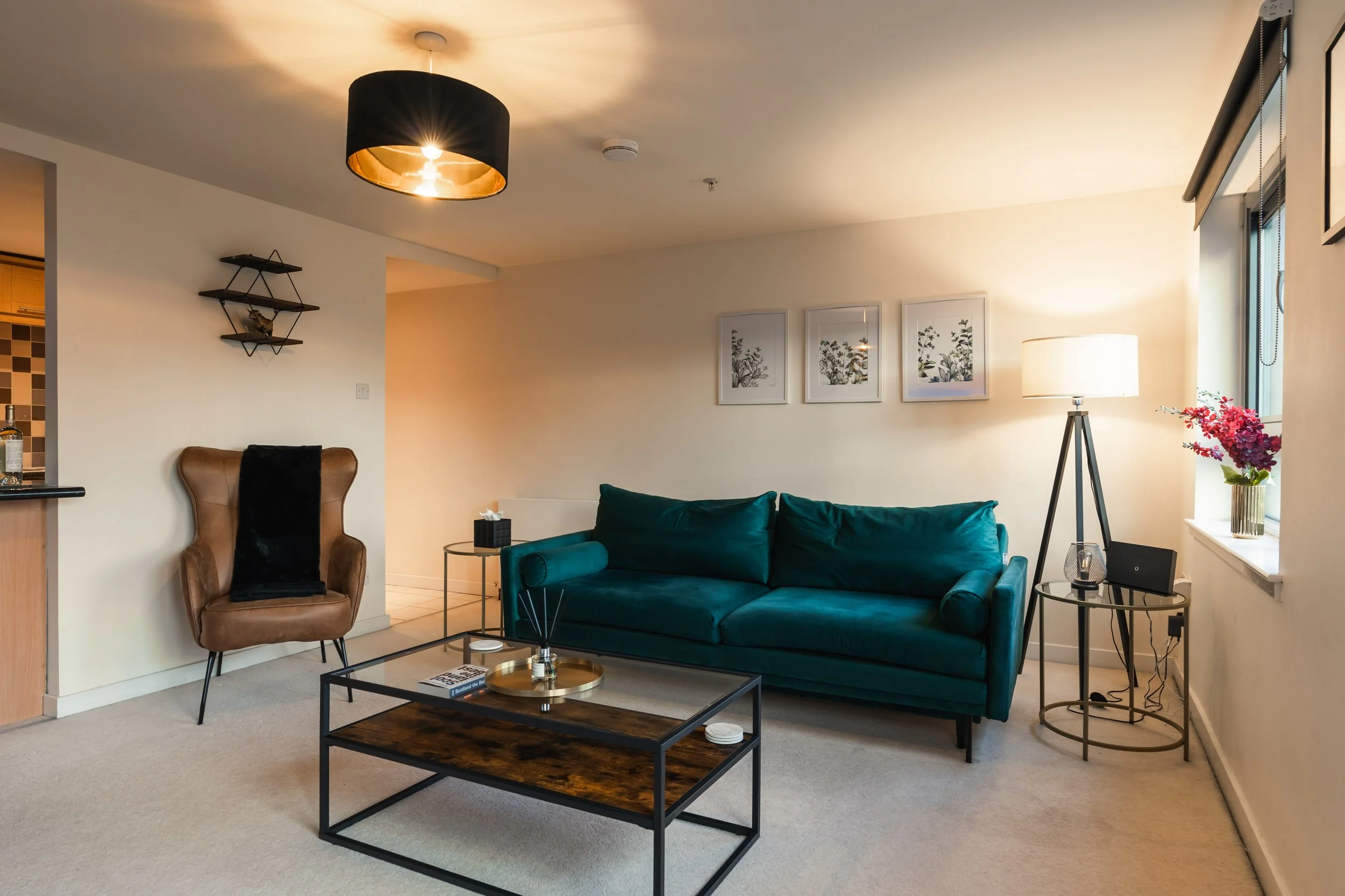 Living room with a teal sofa, a brown armchair, a black coffee table, and a floor lamp near a window with pink flowers on the sill.