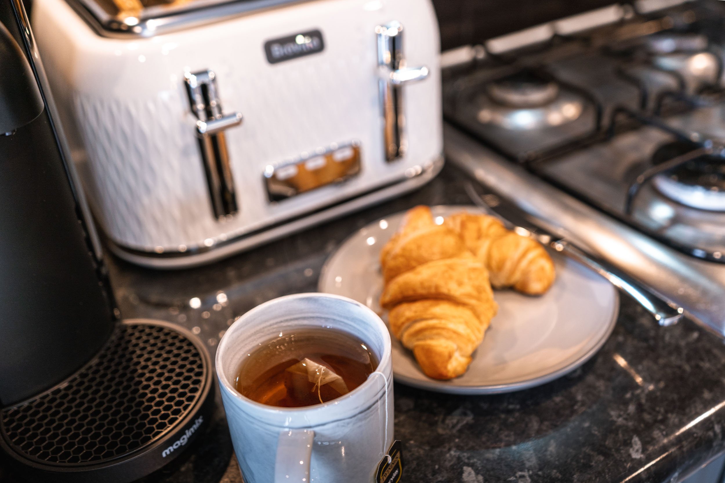 A cup of tea, two croissants on a plate, a toaster, and a stove on a kitchen countertop.