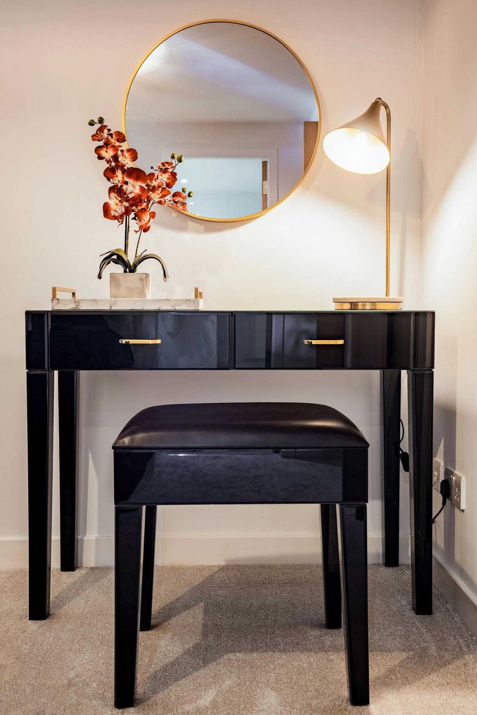 A black vanity table with three drawers and a matching black stool, placed against a white wall. On top of the table, there is a flower vase with red orchid flowers. A round mirror is hung on the wall above the table, and to the right, there is a mod