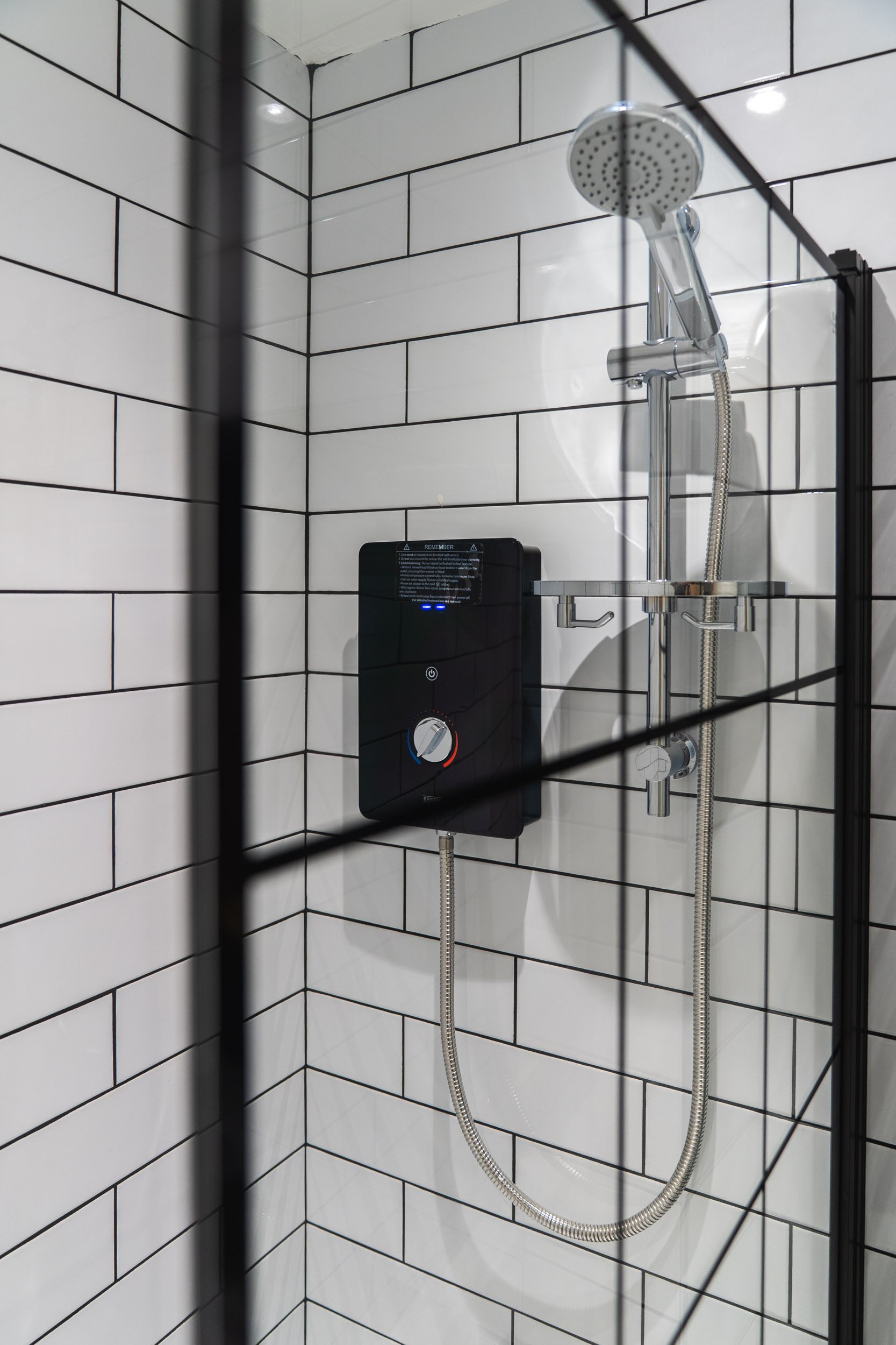 A shower with white subway tile walls, a round showerhead, and black wall-mounted control panel with a dial and digital buttons.