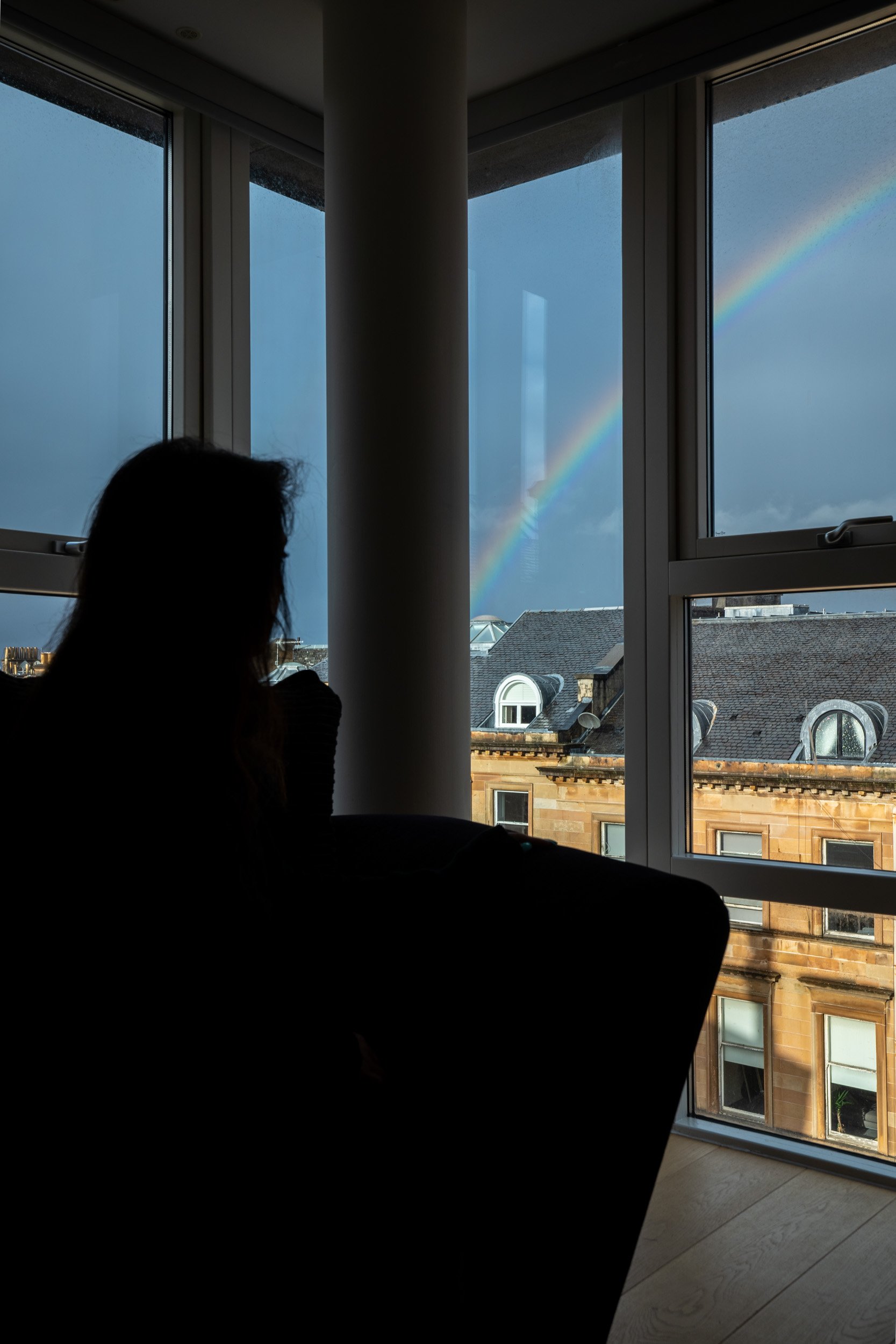 Silhouette of a woman sitting by a large window, looking outside at a rainbow in a stormy sky over a cityscape with old buildings.