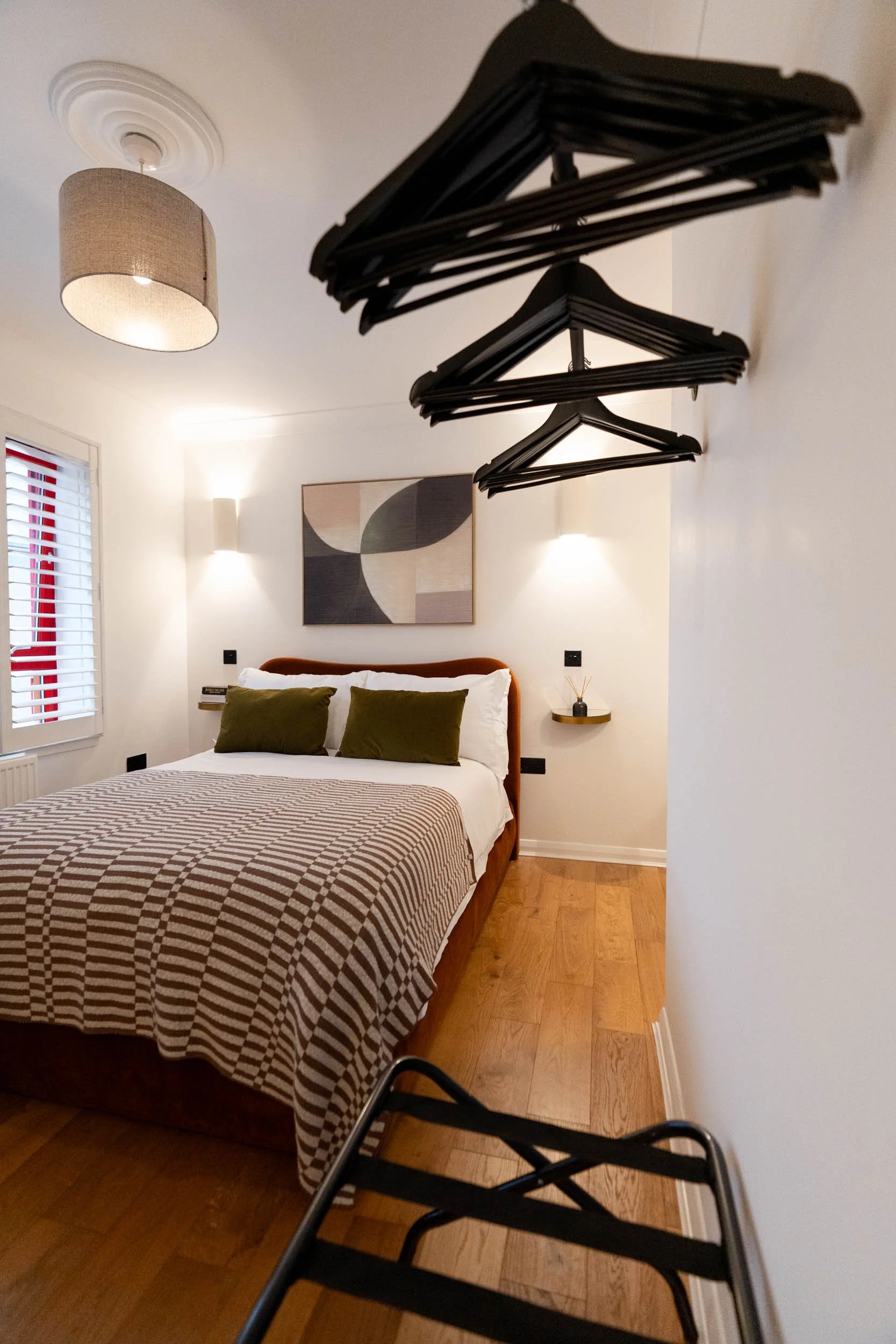 A bedroom with a bed, wall art, two wall lights, and a ceiling light. Hangers are hanging from a wall-mounted rack in the foreground.
