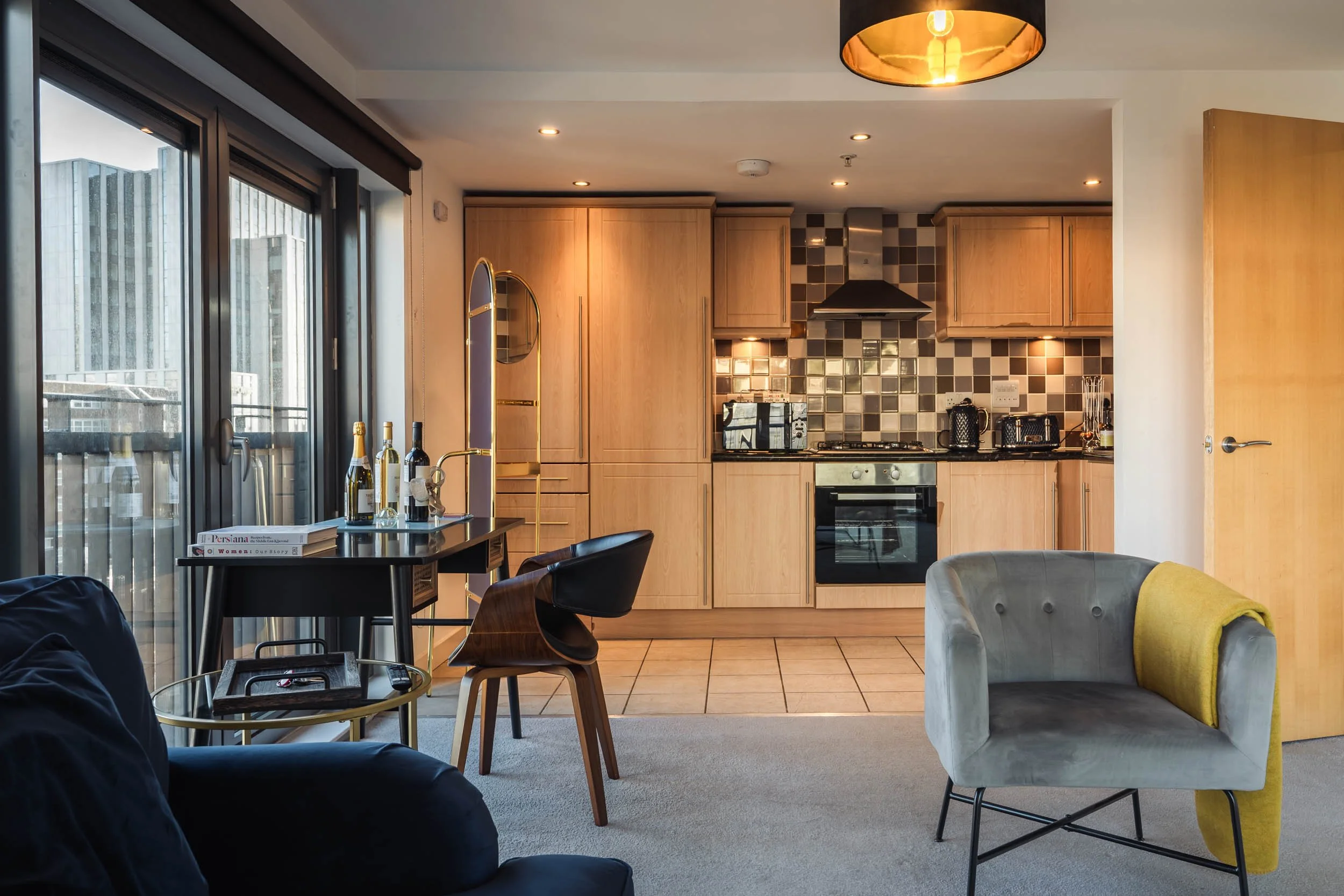 Modern open-plan living space with a view of a kitchen featuring wooden cabinets, a tiled backsplash, and stainless steel appliances. The room has a sliding glass door leading to a balcony, and there is a small dining table with chairs, along with a 