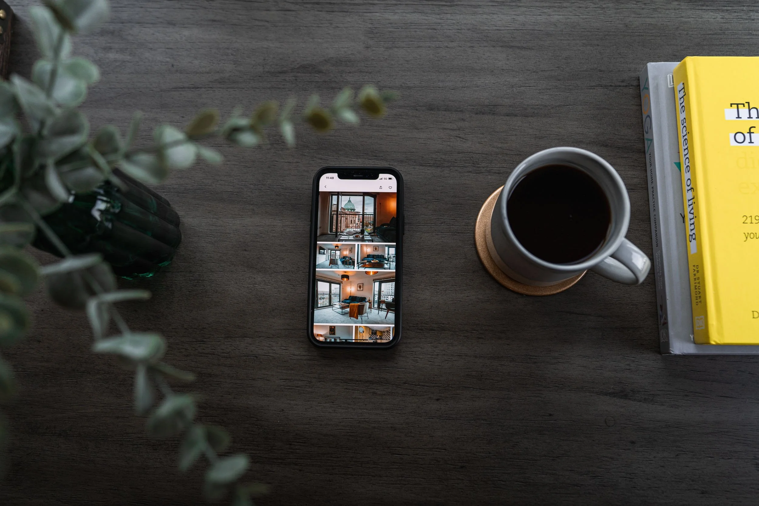 A wooden desk with a smartphone displaying interior photos, a cup of black coffee on a coaster, a stack of books including titles on the science of living, and a potted plant with green leaves.