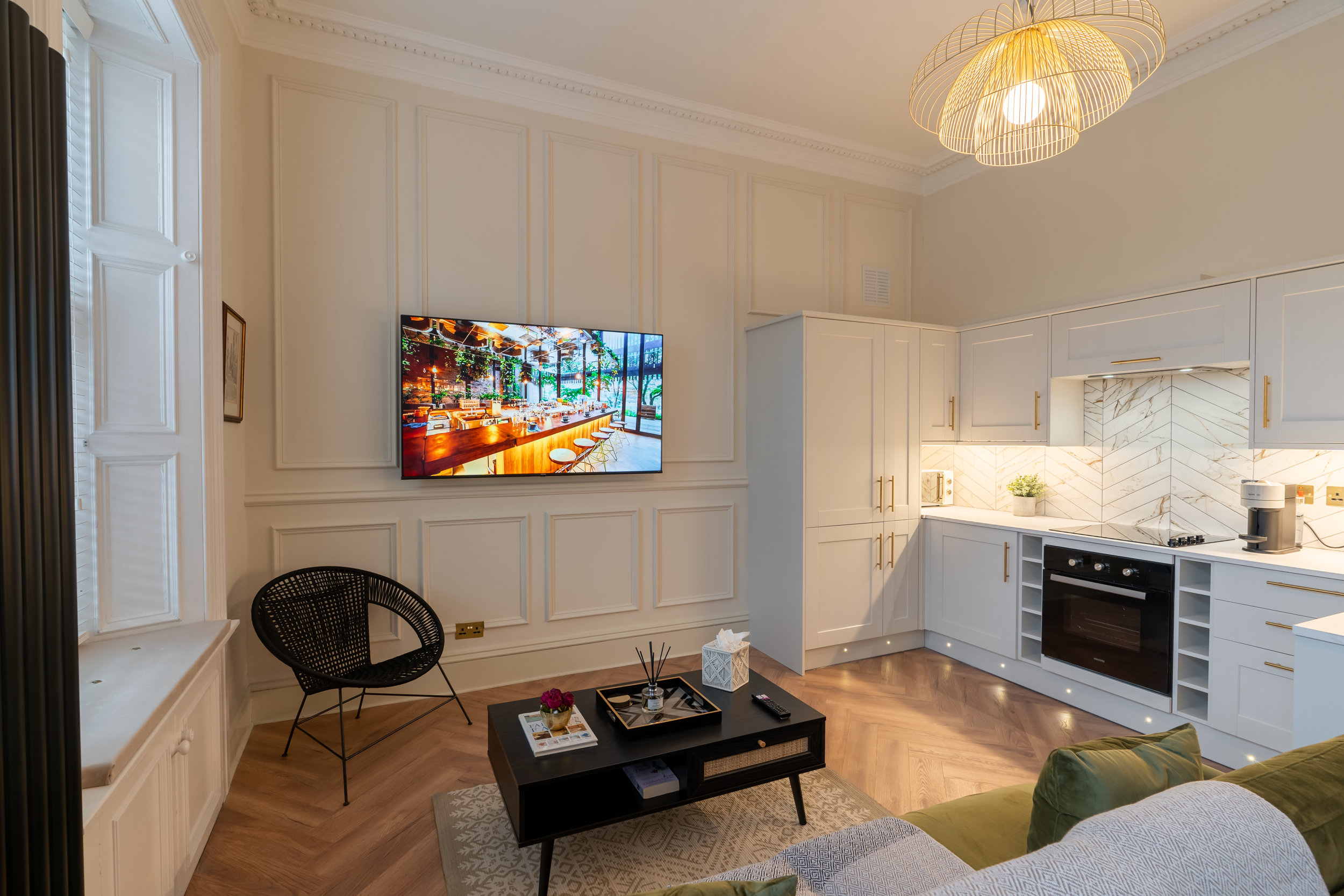 Living room with white kitchen cabinets, a black chair, a coffee table with decorative items, a flat-screen TV on the wall, a green sofa, and a window with shutters.