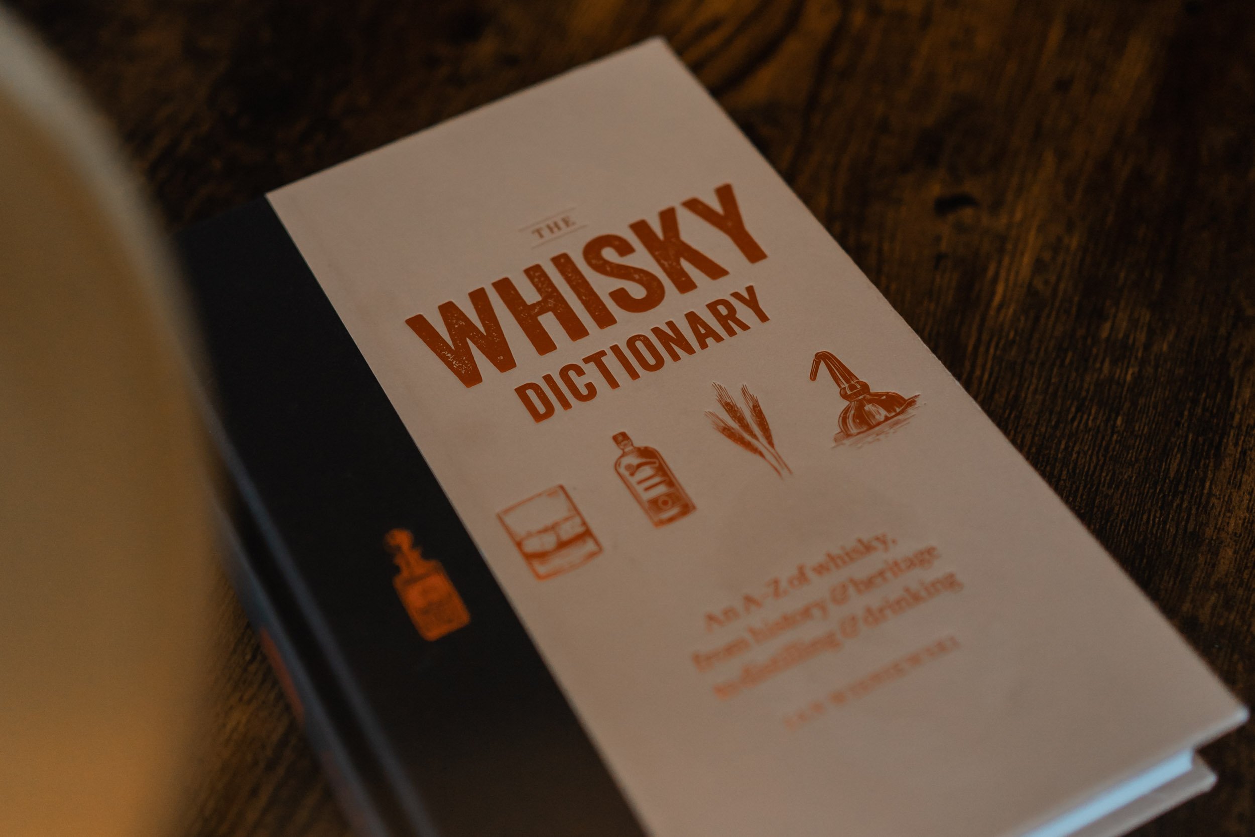 Cover of a book titled 'The Whisky Dictionary' with illustrations of a whiskey glass, whiskey bottle, wheat stalks, and a whiskey still, placed on a wooden surface.