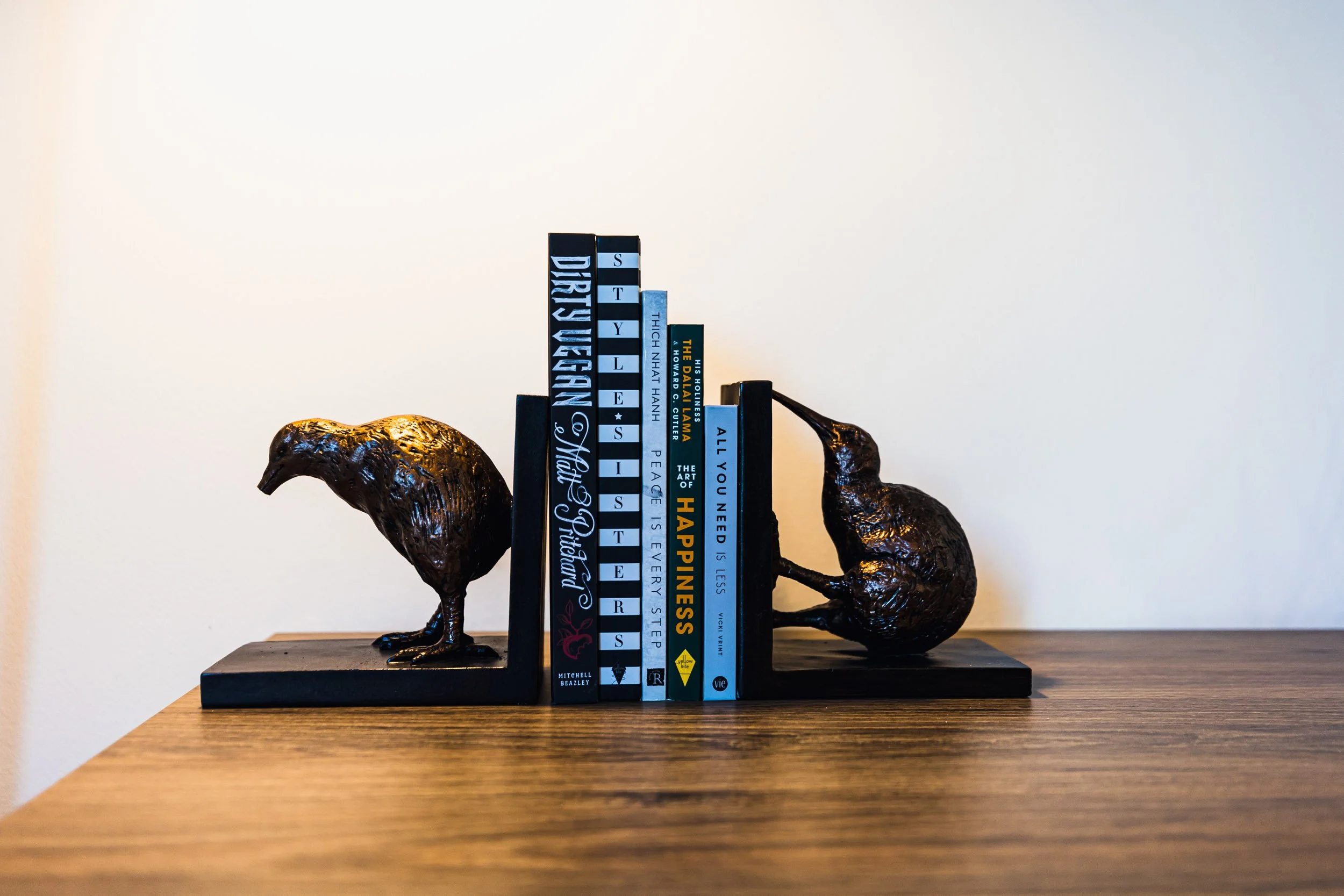 Decorative bookend sculpture of two black birds with textured surfaces, holding a row of colorful books on a wooden surface against a plain wall.