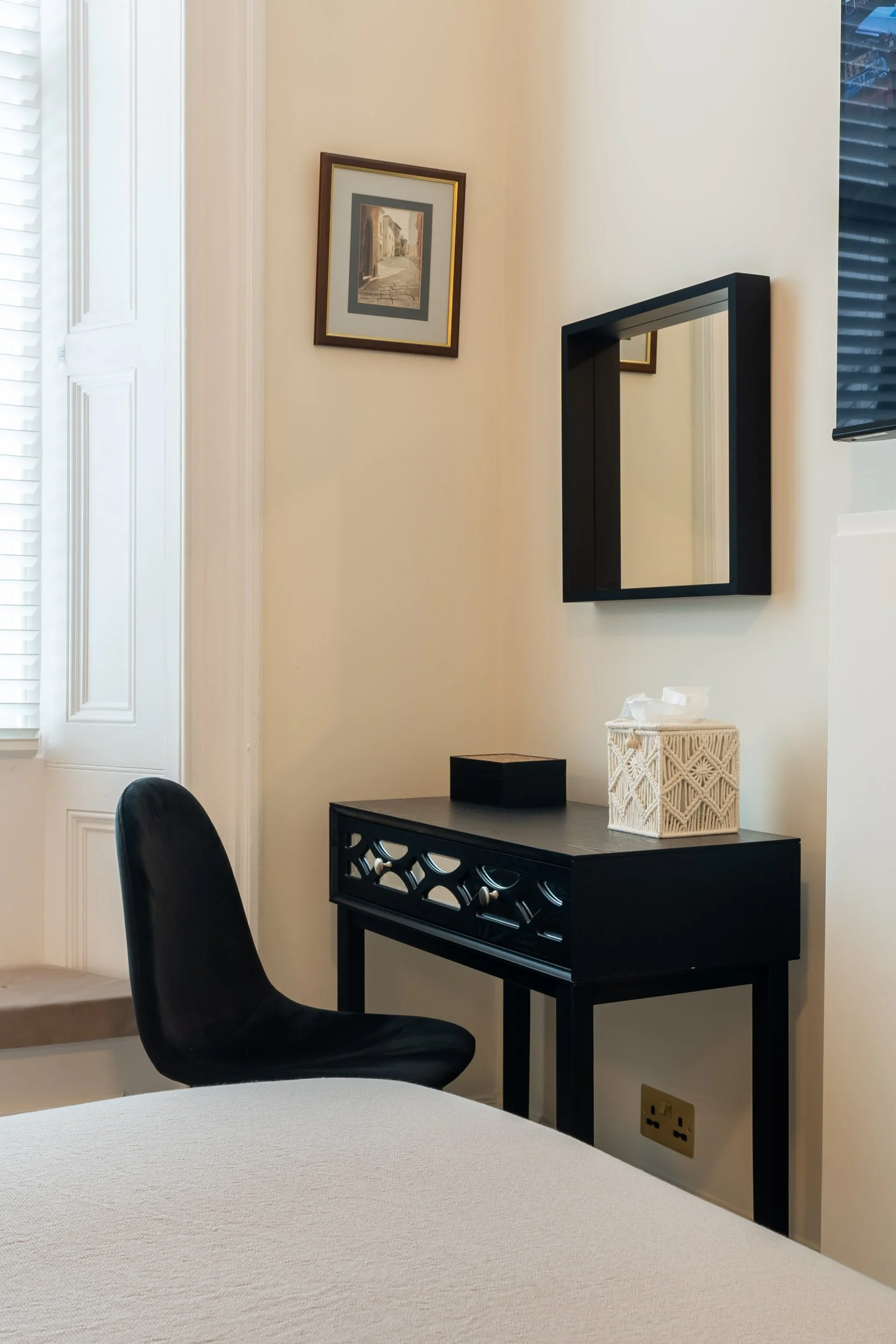 A corner of a room showing a black chair, a black table with decorative cutouts, a tissue box, a small black box, a mirror, a framed painting on the wall, and a window with blinds.