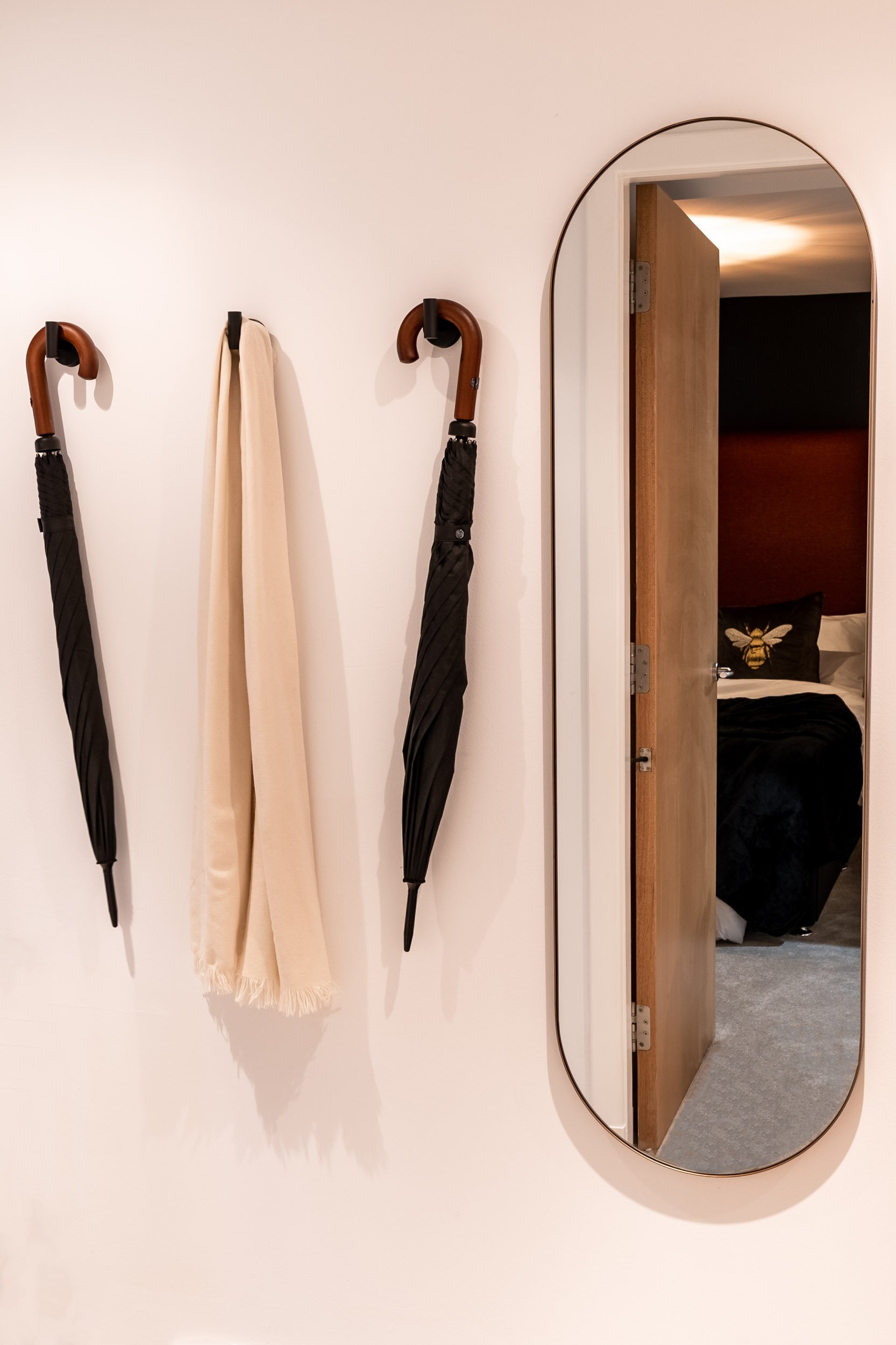 Wall with three umbrellas and a scarf hanging on hooks, with a mirror reflecting a door and a bedroom with a bed and decorative pillows.