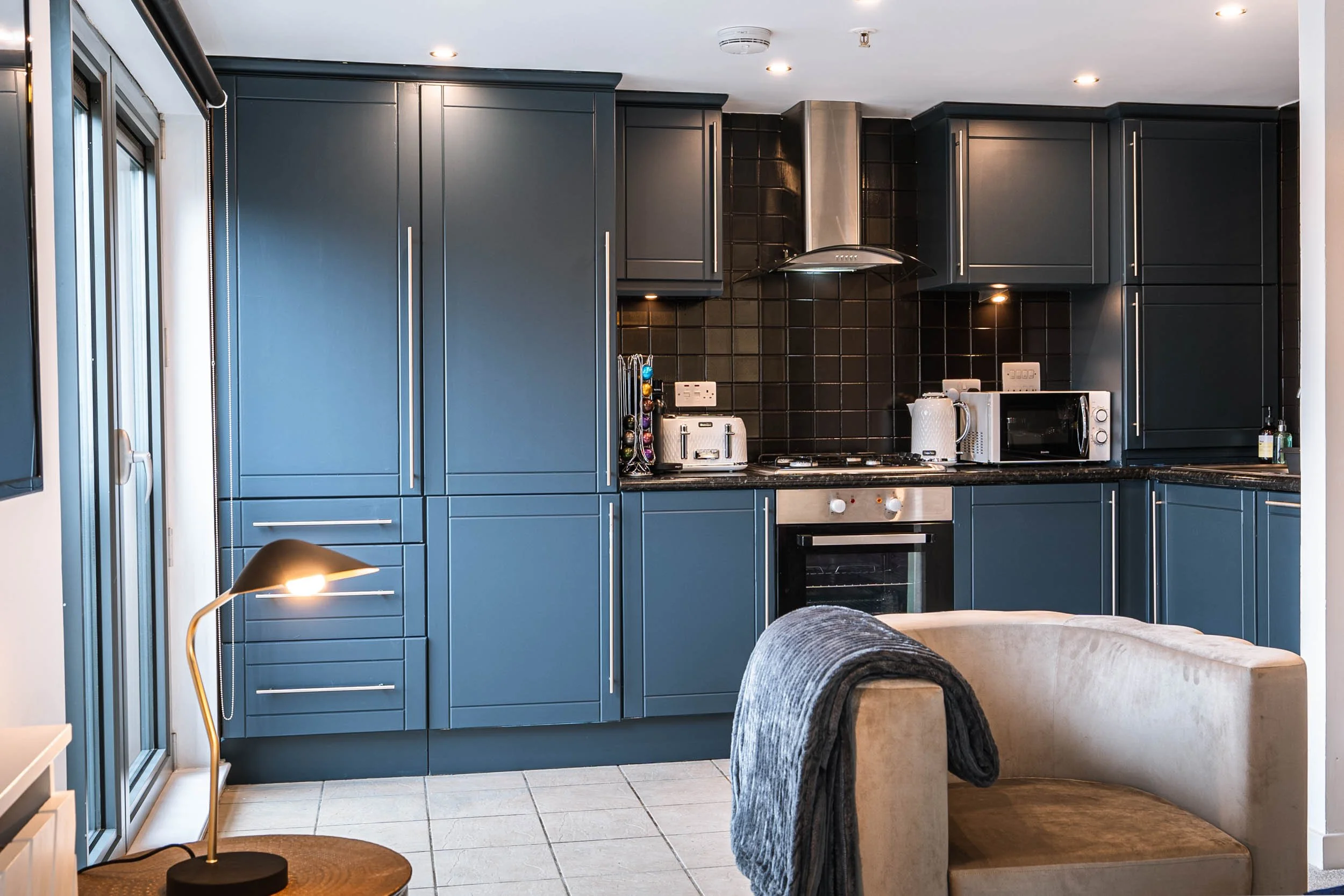 Modern kitchen with blue cabinets, black tiled backsplash, and stainless steel appliances, including microwave, oven, and range hood. Front corner features a beige sofa with a dark gray blanket draped over the arm, and a small wooden side table with 