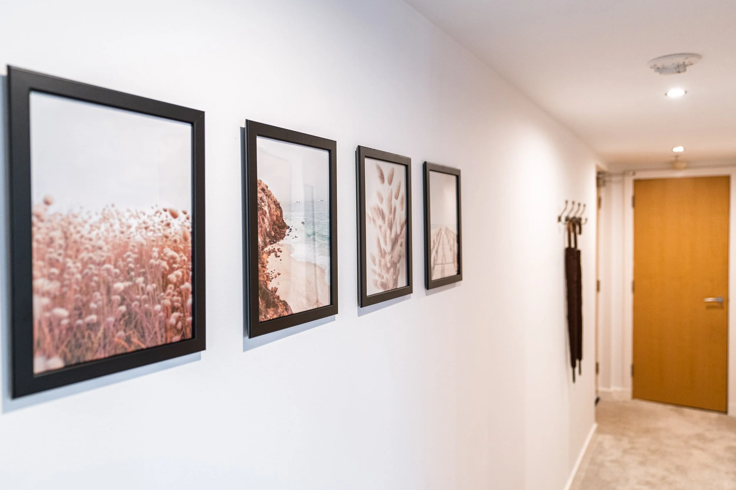 Hallway with five framed landscape pictures on a white wall, with a wooden door at the end and a coat rack on the side.