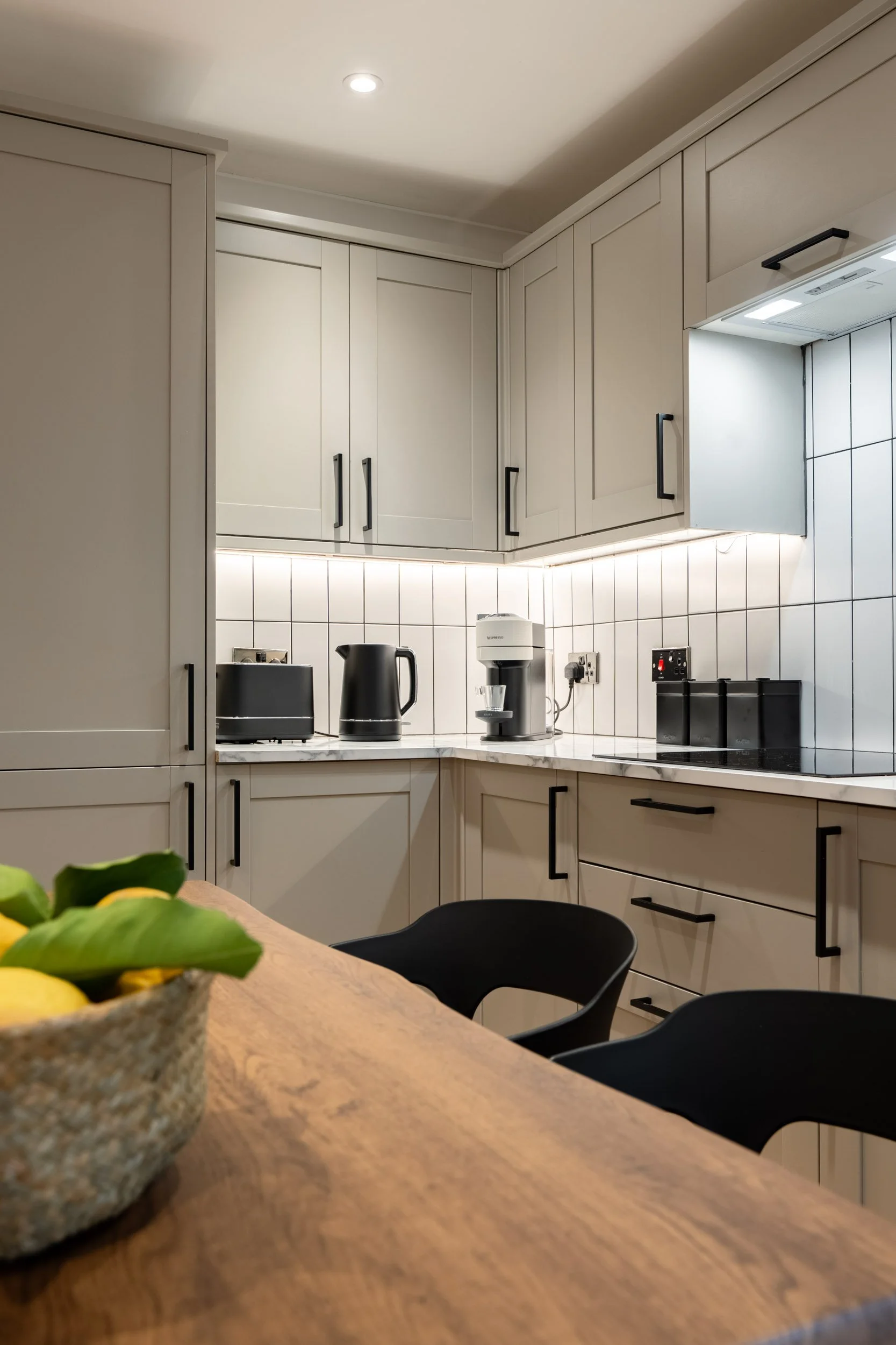 A modern kitchen with white cabinets, black handles, and white tiled backsplash. Countertop appliances include a black toaster, a black electric kettle, and a white coffee machine. Part of a wooden dining table with a basket of lemons and greenery an