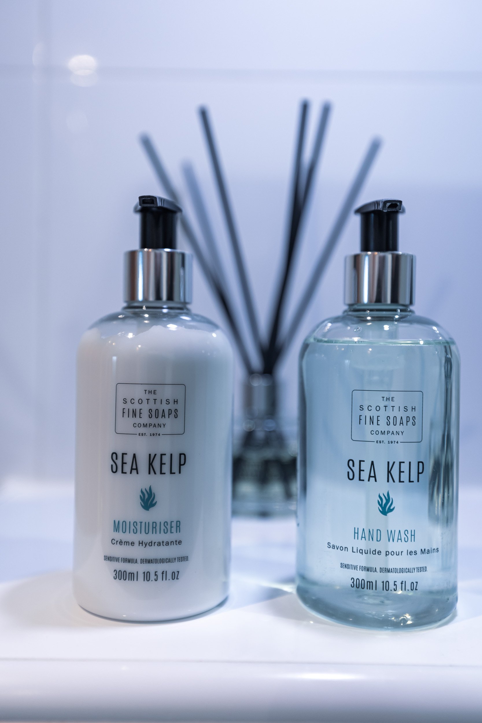 Two bottles labeled 'Sea Kelp' from The Scottish Fine Soaps Company, one as a moisturizer and the other as hand wash, placed on a bathroom shelf with a reed diffuser in the background.