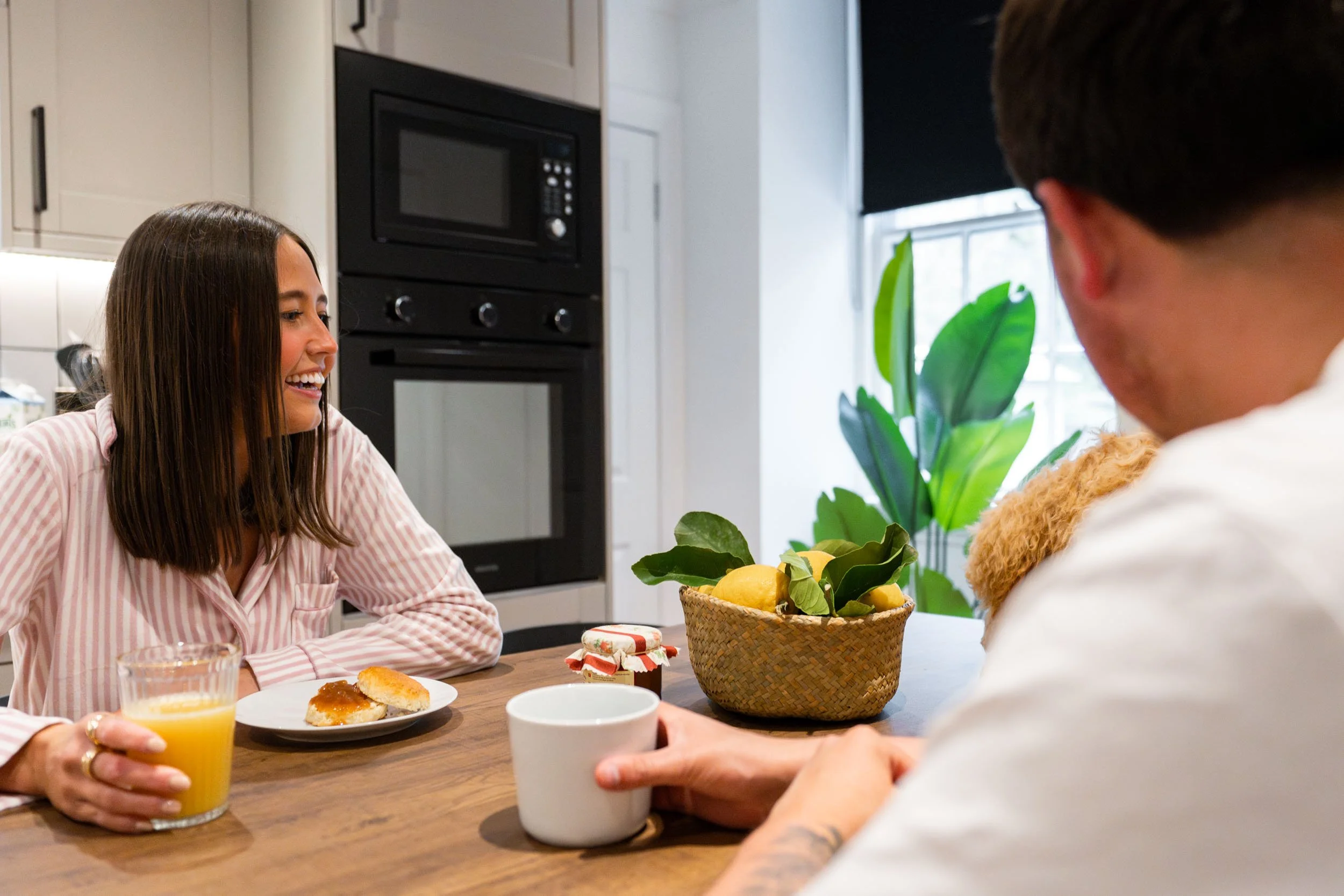 A woman in a pink and white striped pajama top smiling and talking to a man at a kitchen table holding a glass of orange juice. The man, whose face is not visible, is holding a white mug. On the table, there is a plate with two pieces of bread and a 