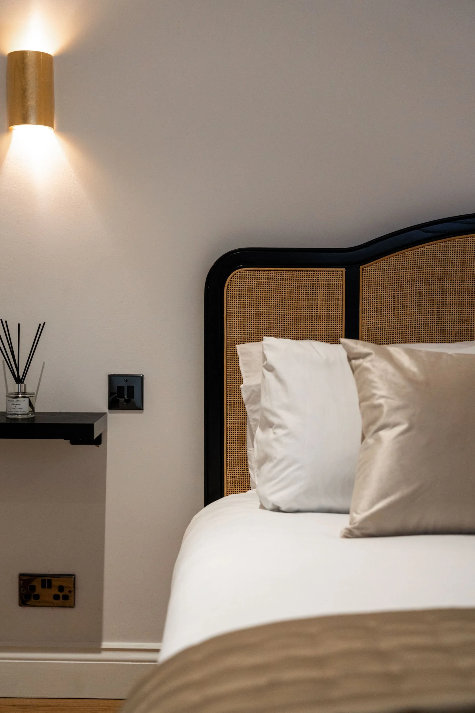 A bedroom with a bed, two pillows, and a headboard with wicker design. A wall-mounted light and a diffuser on a black shelf are visible on a white wall.
