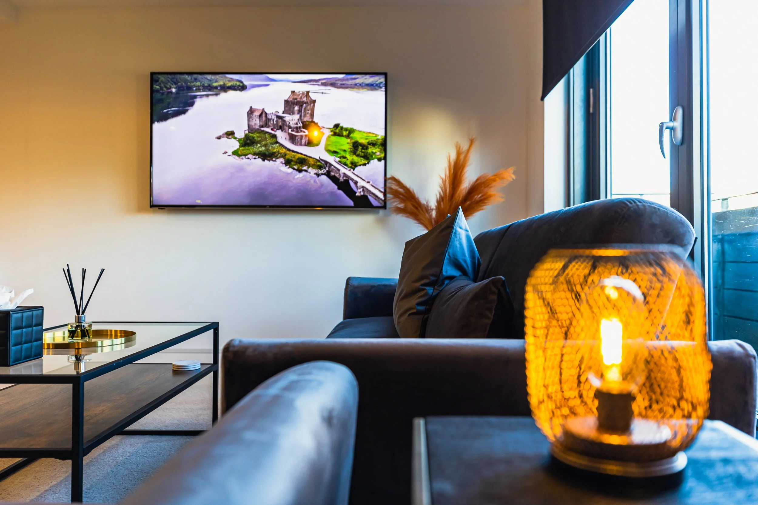 Living room with a wall-mounted TV, a dark-colored sofa with pillows, a glass coffee table, a decorative lamp with an orange glow, and large windows with curtains.