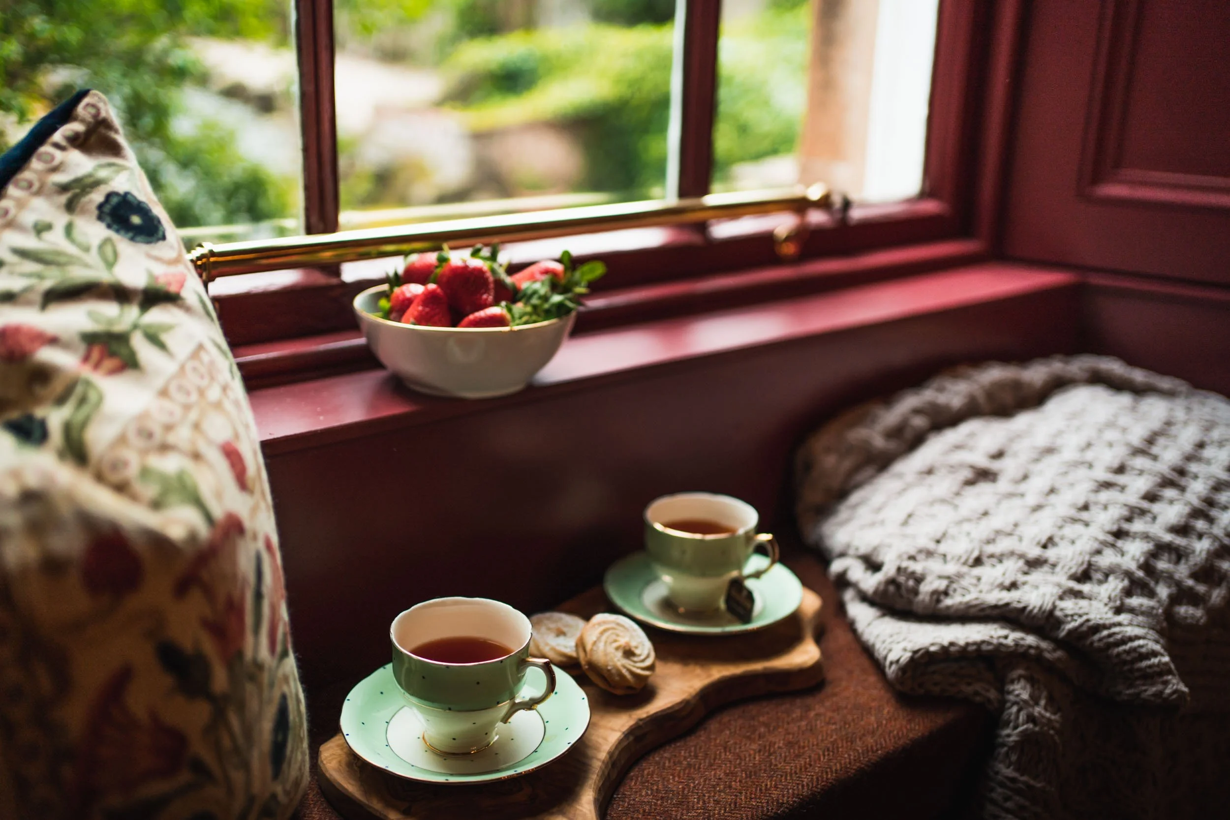 A cozy window seat with two cups of tea, a bowl of strawberries, a pastry, a decorative cushion, a knitted blanket, and a view of greenery outside.