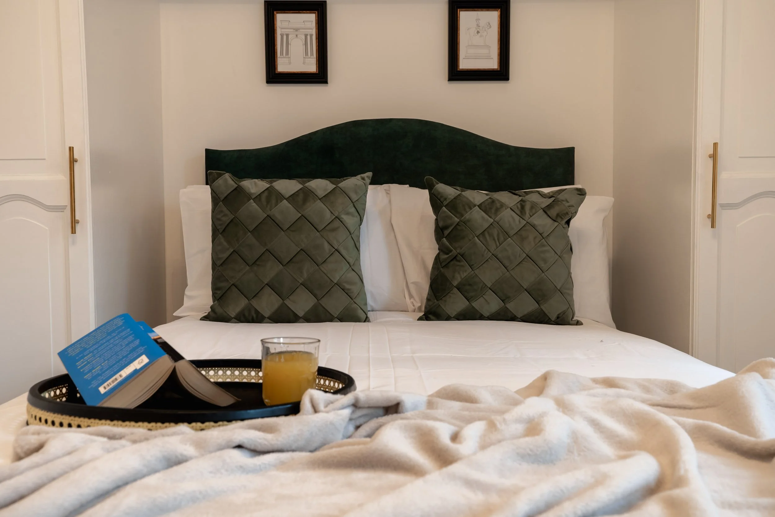 A bed with dark green headboard and two patterned green pillows, set against a white wall with two framed line art pictures. A tray with a book, a glass of orange juice, and a blanket is on the bed.
