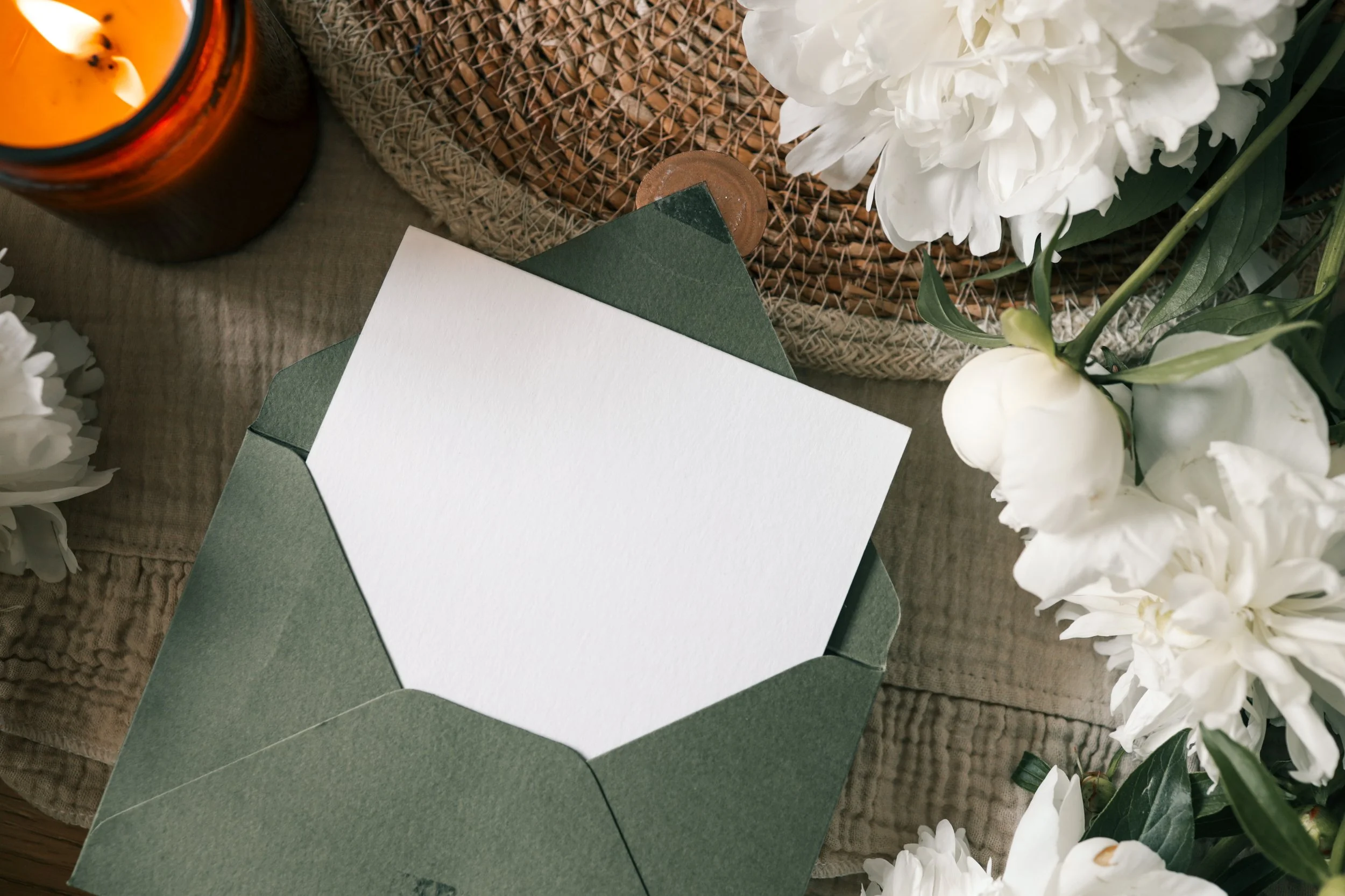 Close-up of a white card and green envelope. Flowers, a candle burning and a wicker mat style the image. 