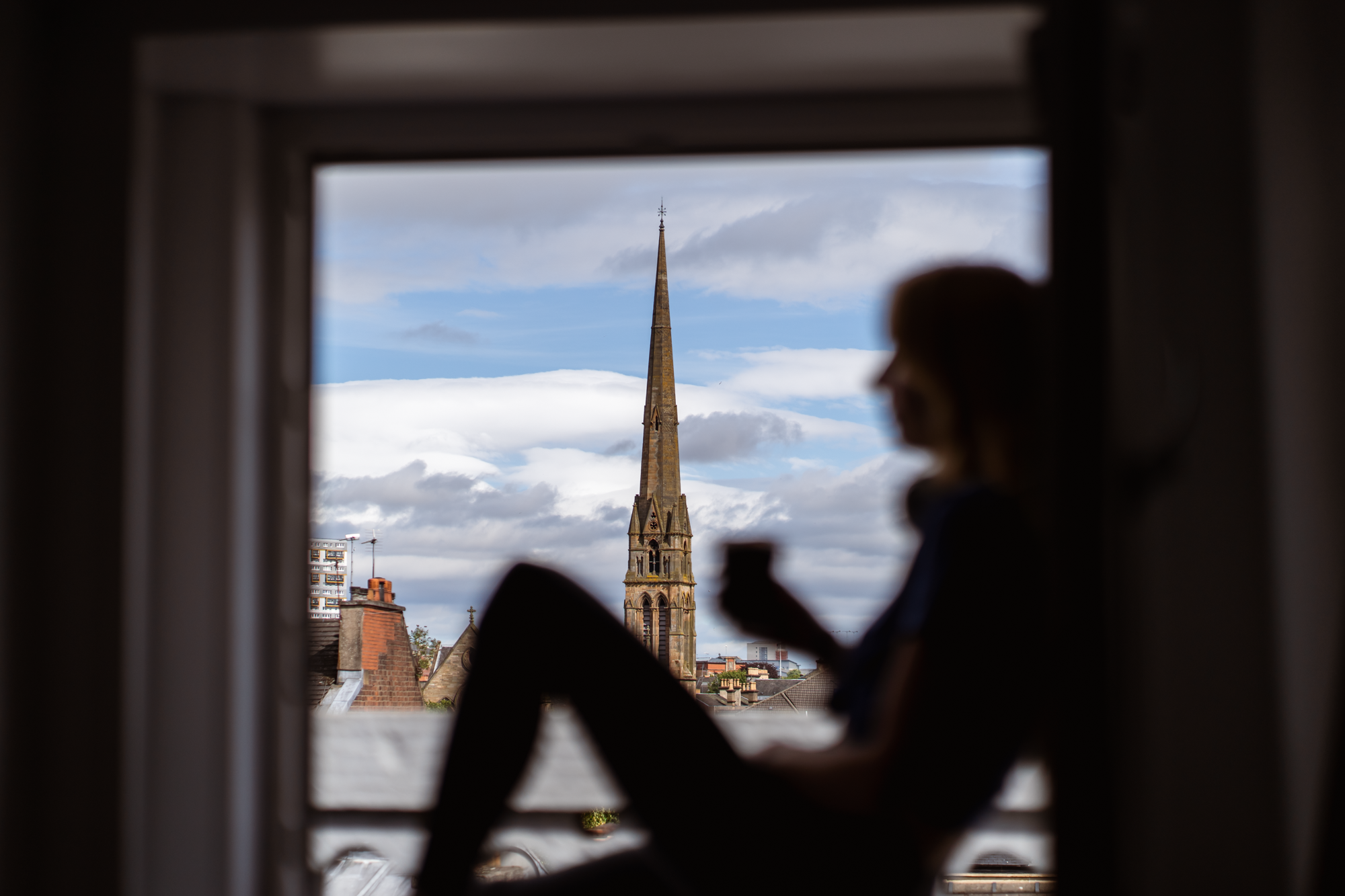 Silhouette of a person sitting on a windowsill with a cityscape and a church steeple in the background