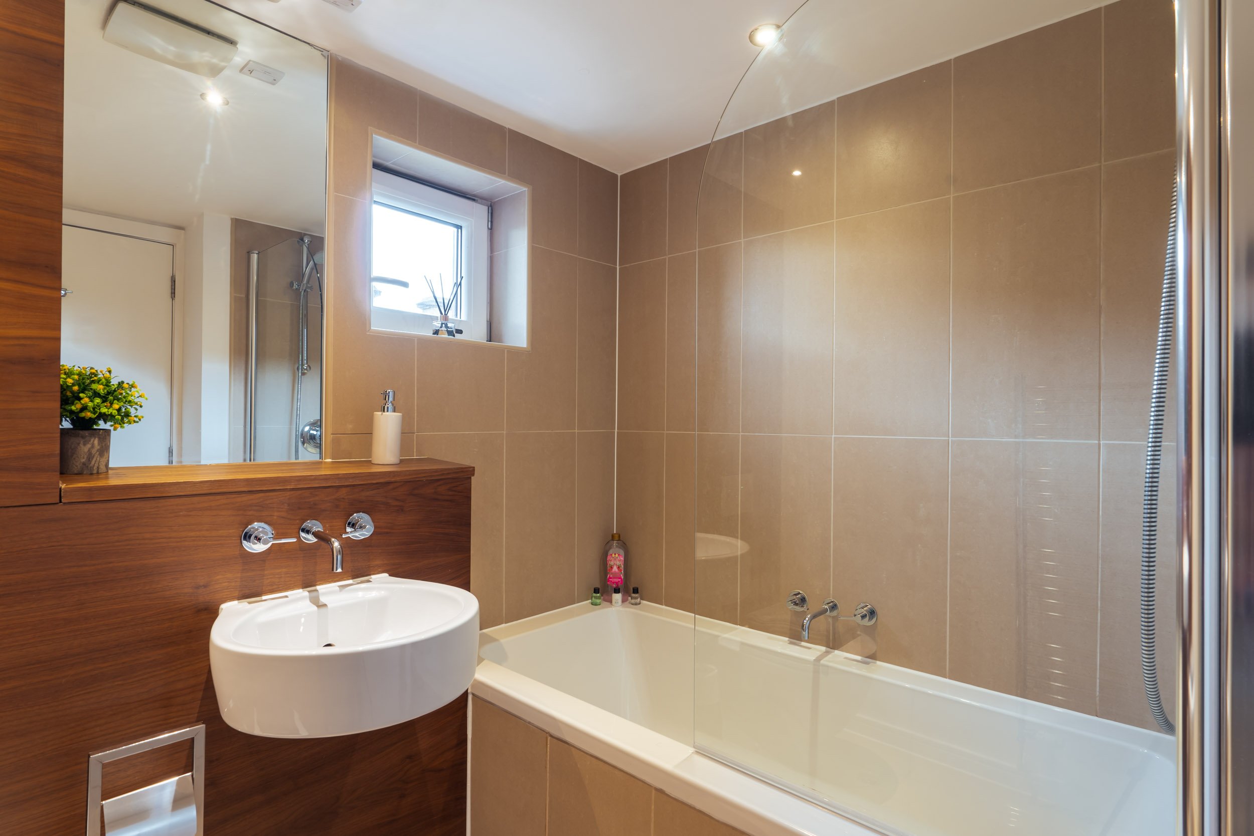 A modern bathroom with beige tiled walls, a small window, a white bathtub with a curved glass shower screen, a white sink with a wooden vanity, a large mirror, a soap dispenser, a plant near the mirror, and some bottles on the corner of the bathtub.