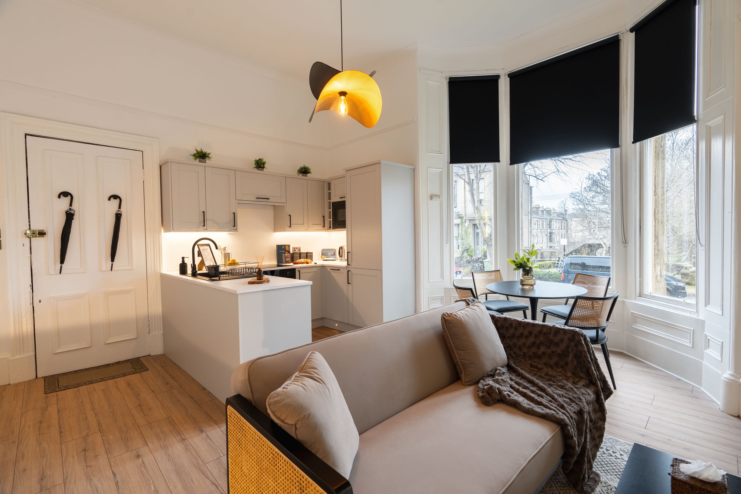 Living room with a beige sofa, four black chairs around a round table near large bay windows with black blinds, a small dining area with plants, white kitchen with gray cabinets, and a modern ceiling light fixture.
