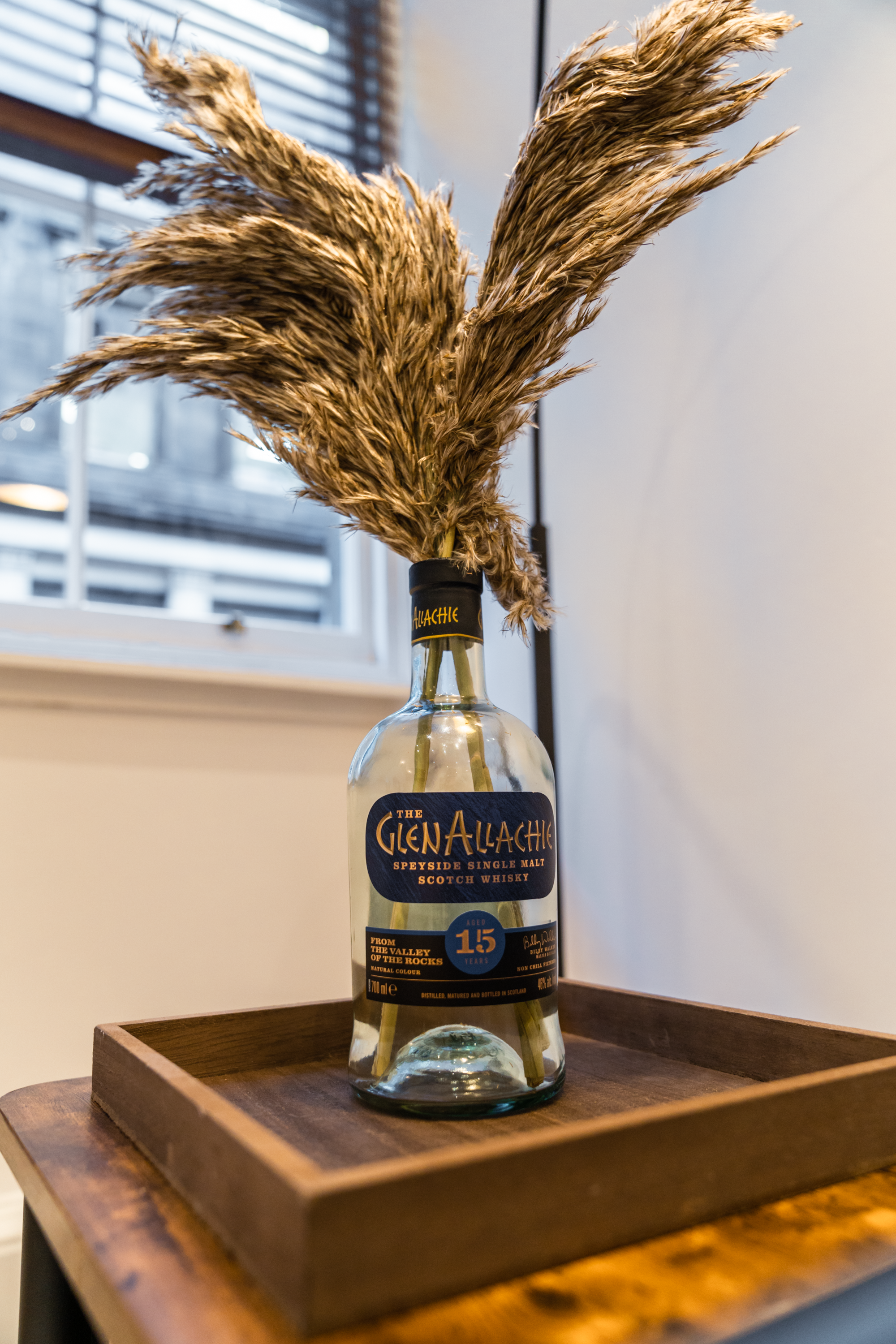 A bottle labeled The Glen Allachie Speyside single malt Scotch whisky with dried pampas grass arranged inside, placed on a wooden tray near a window.