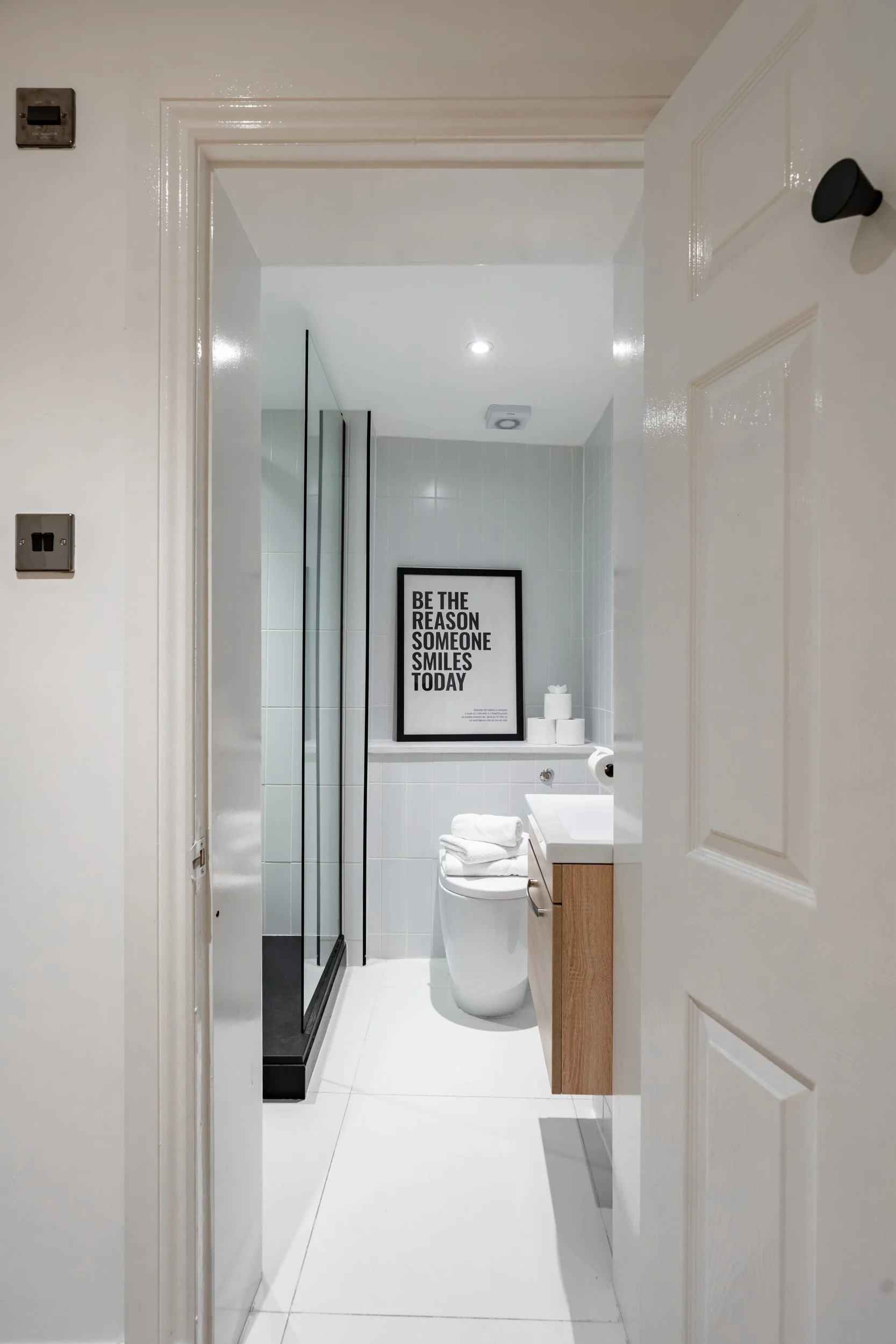 Bathroom with white tiled walls, a framed quote saying "Be the reason someone smiles today," a toilet with folded towels on top, a black framed mirror, a wooden vanity with a sink, a glass shower enclosure, and white toilet paper rolls.