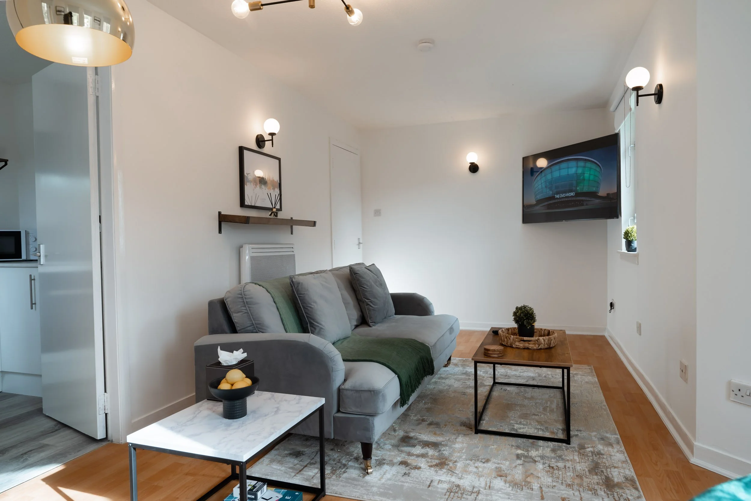 Living room with a gray sofa, a small marble side table with lemons, a wooden coffee table with plants, wall-mounted TV, and various light fixtures on white walls.
