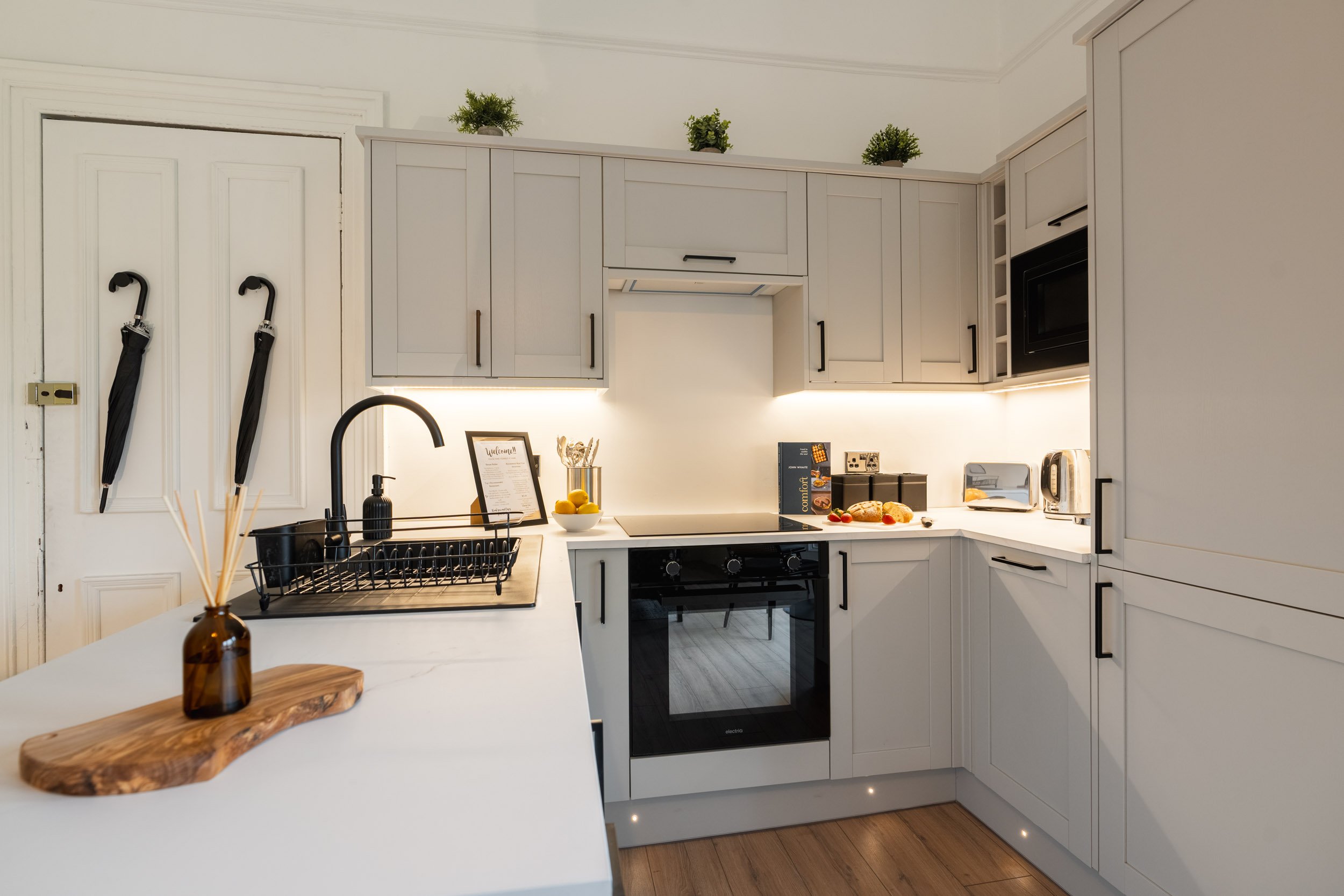 Modern kitchen with white cabinets, black handles, built-in microwave, oven, and small appliances on a white countertop, with green plants on top of cabinets, a black dish rack, and umbrellas hanging on the door.