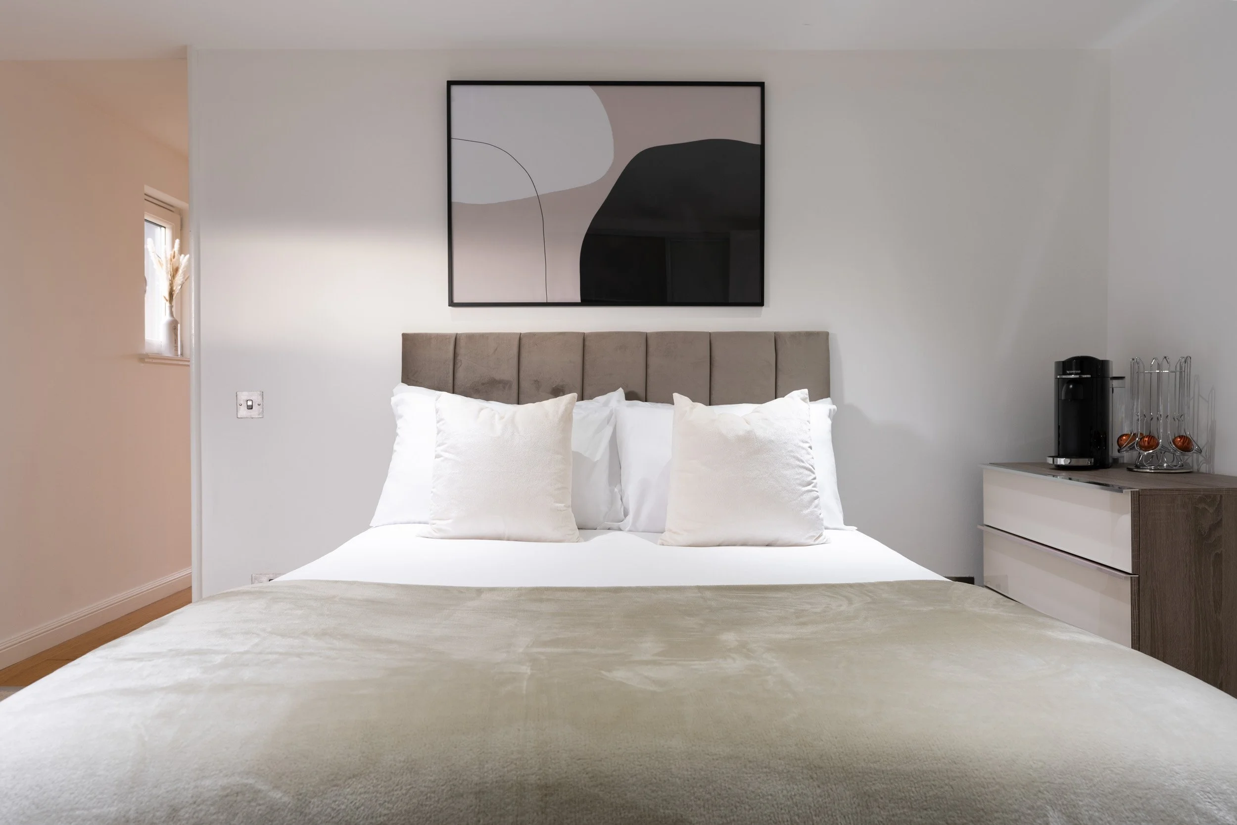 Modern bedroom with a bed, beige headboard, white pillows, abstract artwork above the bed, and a side table with a coffee maker.