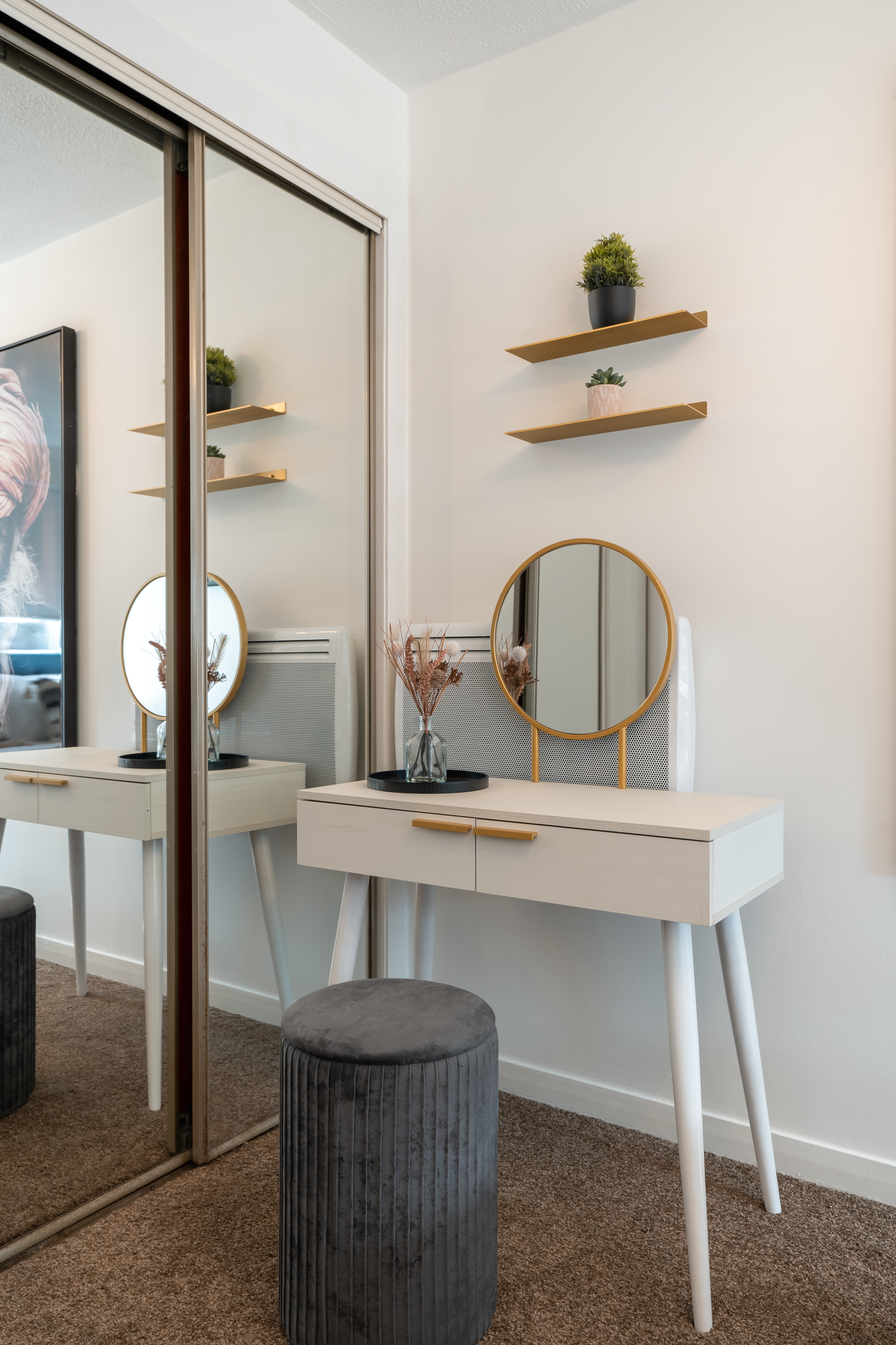 A bedroom corner with a white vanity table, a round mirror, a vase with pink flowers, two black trays, and a black velvet ottoman. Two wooden floating shelves with potted plants are on the wall, and a mirrored closet reflects the scene.